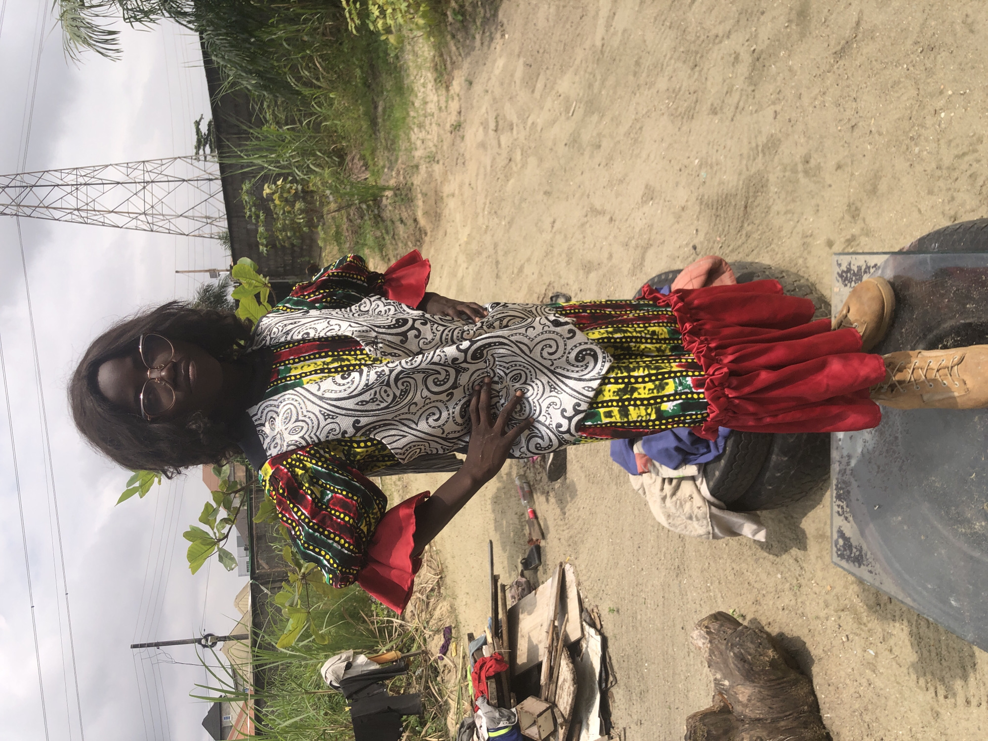 In Ado, Nigeria, a young woman with dark, curly hair and rose-tinted glasses stands on two stacked tires, her hands on her hips. She is wearing a vibrant, multi-colored African print dress with puffy, red-trimmed sleeves and a ruffled hem. The dress features a black and white swirling pattern on the bodice. Behind her, the ground is sandy and appears to be a dirt lot. To the left, there's a pile of wooden planks and debris, with some clothing items hanging on a rack in the background. Further back, tall green grass and bushes line a wall, with a large power pylon visible against a cloudy sky. The overall scene suggests an outdoor setting in a semi-urban or rural area.