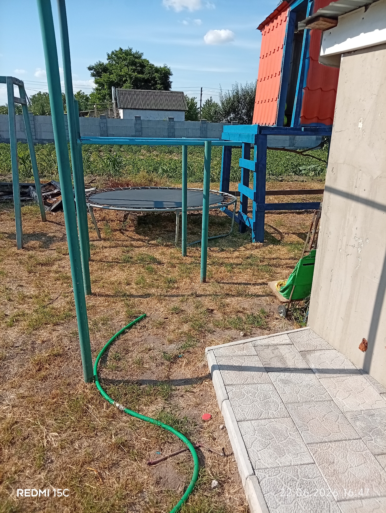 The image depicts an outdoor scene in Mykolaivka, Ukraine, likely a backyard or recreational area, under a bright blue sky with scattered white clouds. In the foreground, a green garden hose lies coiled on dry, yellowish grass. A grey concrete patio with textured tiles is visible to the right. Behind this, a trampoline with a black mat is set up, supported by a metal frame. Adjacent to the trampoline, a blue metal structure with orange tiled roofing rises, appearing to be part of a playhouse or fort. A grey concrete wall or fence runs across the middle ground, with trees and a small building visible beyond it. A green utility bag rests on a small wheeled cart near the structure. The overall impression is one of a slightly overgrown, lived-in outdoor space.