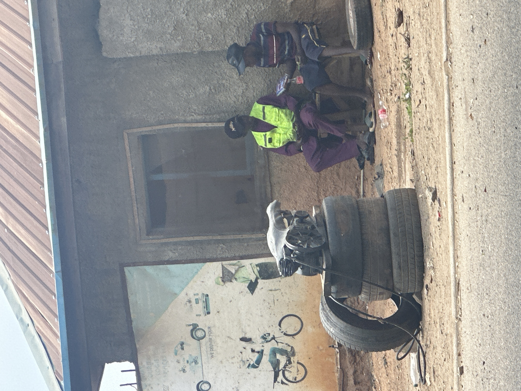 Two men sit near a building in Abuja, Nigeria. One man wears a yellow vest and purple clothing. The other man wears a striped shirt and dark pants. Tires are stacked near the building, and an engine sits on top of the tires. A sign on the building depicts vehicles, including a bicycle and van. The area appears to be a roadside or mechanic shop.