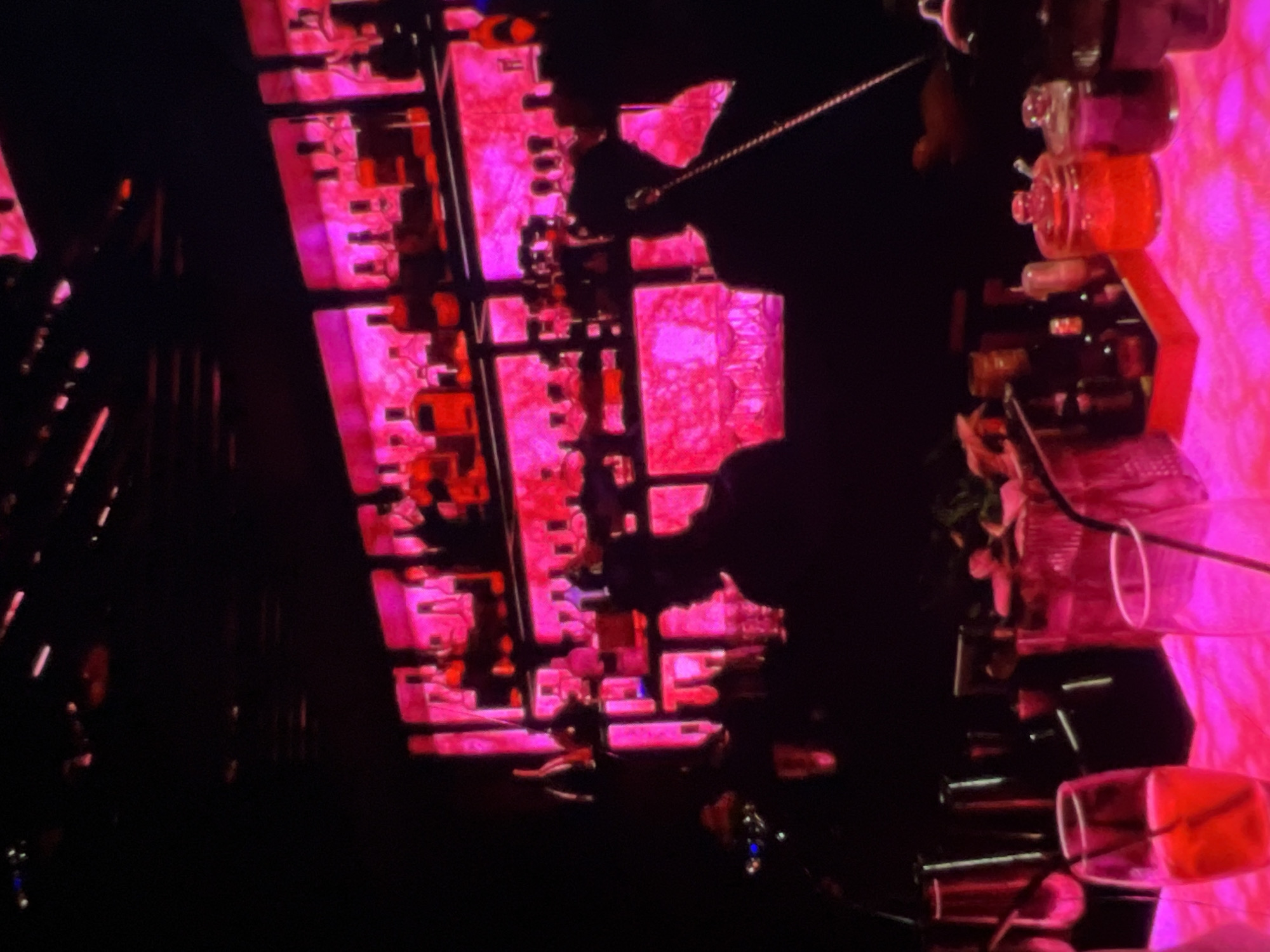 The image depicts a dimly lit bar scene, likely in Lagos, Nigeria, illuminated by vibrant pink and red neon lights. The background features shelves stocked with numerous bottles of alcohol, glowing brightly. In the foreground, two figures are silhouetted behind a bar counter. One figure, on the right, is wearing headphones and appears to be working, possibly a bartender. The other figure on the left is less distinct. The bar counter itself is covered in a textured pink surface, with several glasses and cocktail ingredients visible. One glass in the lower left corner contains a red liquid and a straw. The overall atmosphere is that of a lively nightlife establishment.