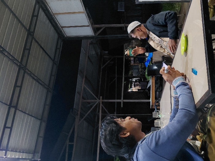 A low-angle shot shows two men at a table. One man, wearing a white skullcap, a black jacket, and a patterned scarf, gestures with his hands. The other man, in a blue sweatshirt, holds a smartphone and appears to be filming or photographing. A green plate sits on the table near a lighter. The setting appears to be an outdoor or semi-outdoor space with a corrugated metal roof. It is nighttime, and the area is illuminated by artificial lighting.