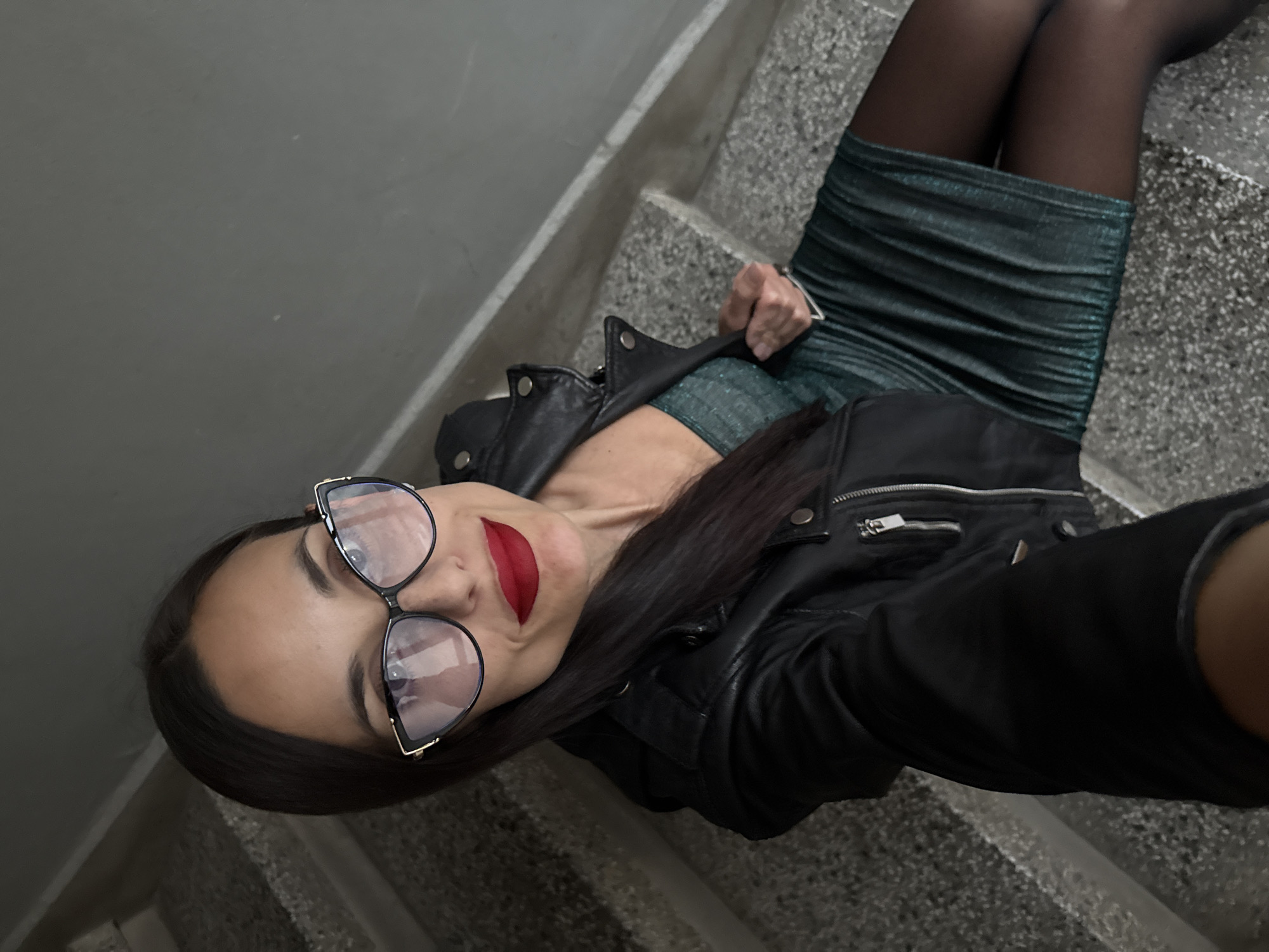 A woman with long, dark hair, wearing large glasses, a black leather jacket, a dark green fitted dress, and black tights, is lying on a set of concrete stairs. She is wearing red lipstick and is looking at the camera. The background consists of the concrete stairs and a light gray wall. Location information for Pleven, Bulgaria is not available in the image.