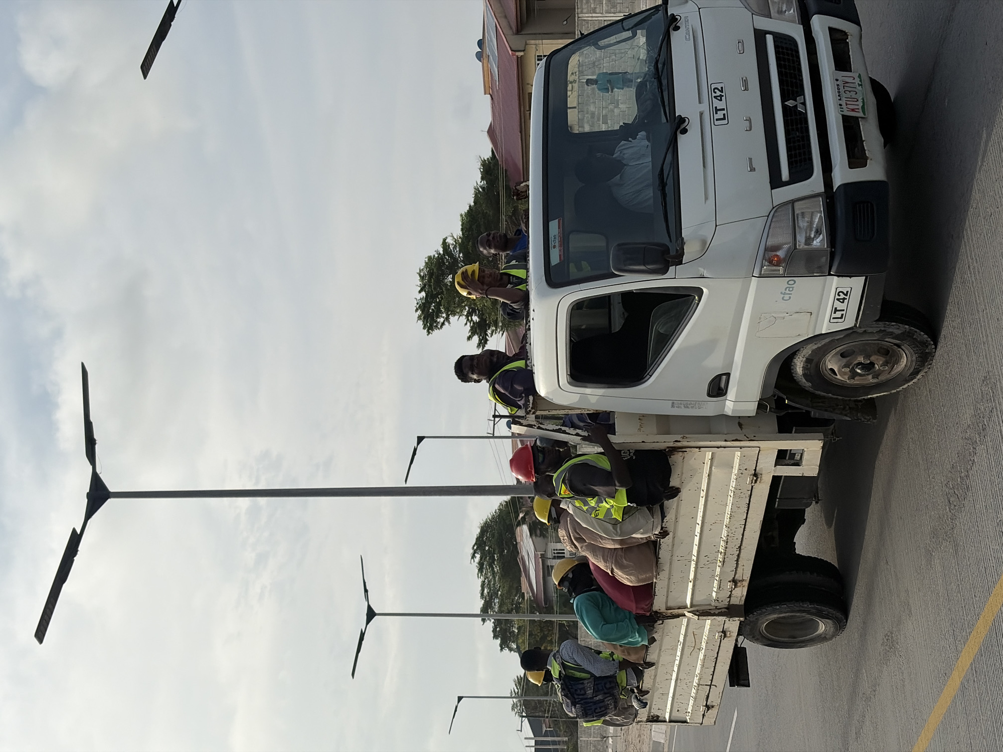 A white FUSO CANTER flatbed truck is driving on a paved road with yellow lane markings in Alaguntan, Nigeria. The truck is carrying approximately ten individuals in the back, some wearing hard hats and reflective vests. They appear to be construction workers. The truck's license plate is visible as KTU 37YJ. In the background, there are houses with red roofs and streetlights. The sky is overcast.