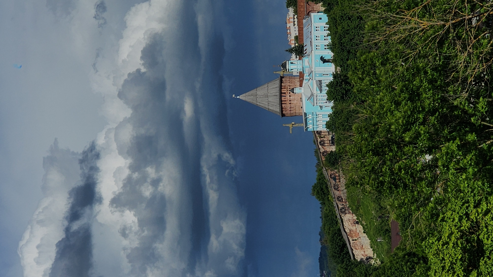The image shows a view of a historic complex against a sky filled with dramatic clouds. A light blue building with white trim and a brown-topped tower stands prominently in the center. The architecture is characterized by a tall, conical roof on the tower, which is connected to a larger, ornate building. The structure appears to be part of a larger complex that may be either a religious or civic monument. Lush green trees and vegetation surround the structure, particularly on the right side. Below the building, a section of a brick wall is visible. The sky is the dominant element, featuring a mix of dark storm clouds and patches of lighter, cumulus clouds. The overall perspective suggests the shot was taken from a lower vantage point. The location is Unknown, Unknown.