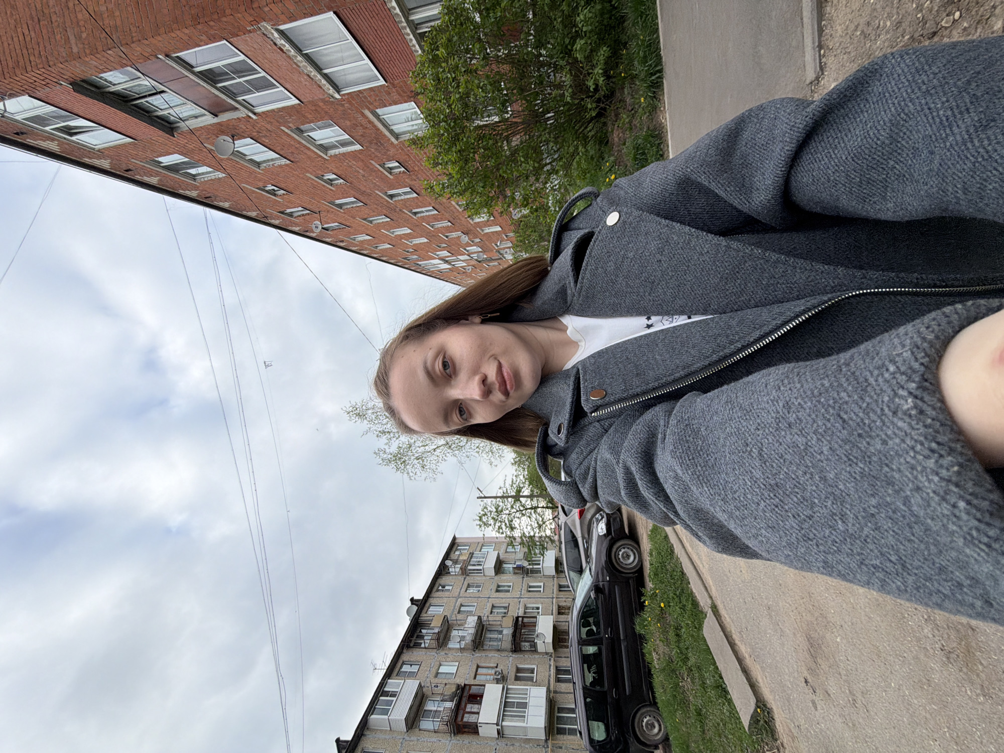 A woman is taking a selfie outdoors. She's wearing a grey coat over a white shirt. In the background, there are multi-story apartment buildings and a cloudy sky. A black car is parked along the sidewalk. The location is identified as Reshetnikovo, Russia.