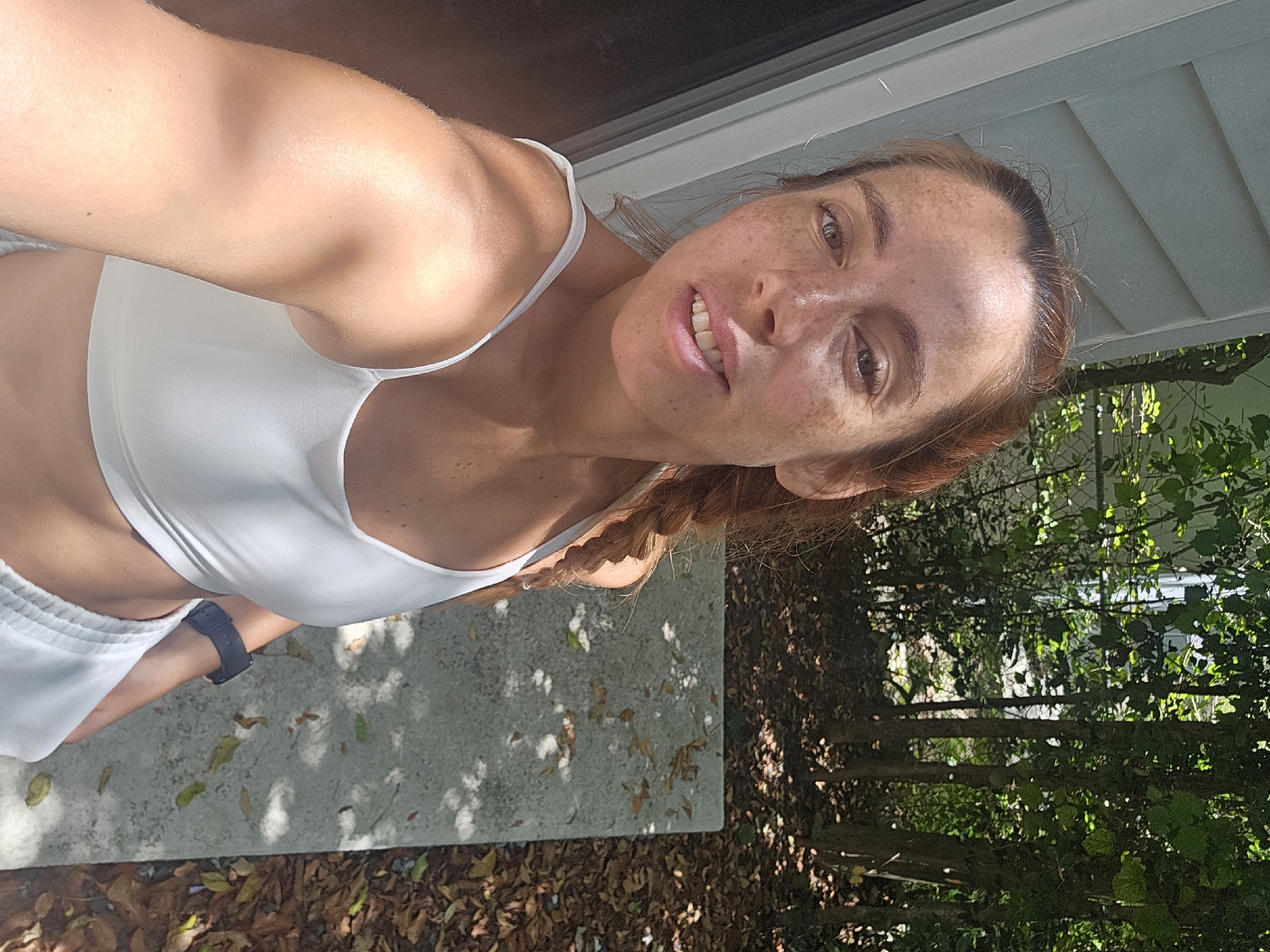 A young, fair-skinned woman with red hair, a braided ponytail, and freckles is taking a selfie outdoors in Islamorada, United States of America. She is wearing a white sports bra and white sweatpants. Her left arm is extended towards the camera, and she is looking directly at it with a slight smile. The background shows a residential building with light-colored siding, trees with green foliage, and fallen leaves on a concrete path. The sun is shining, casting shadows on the ground.