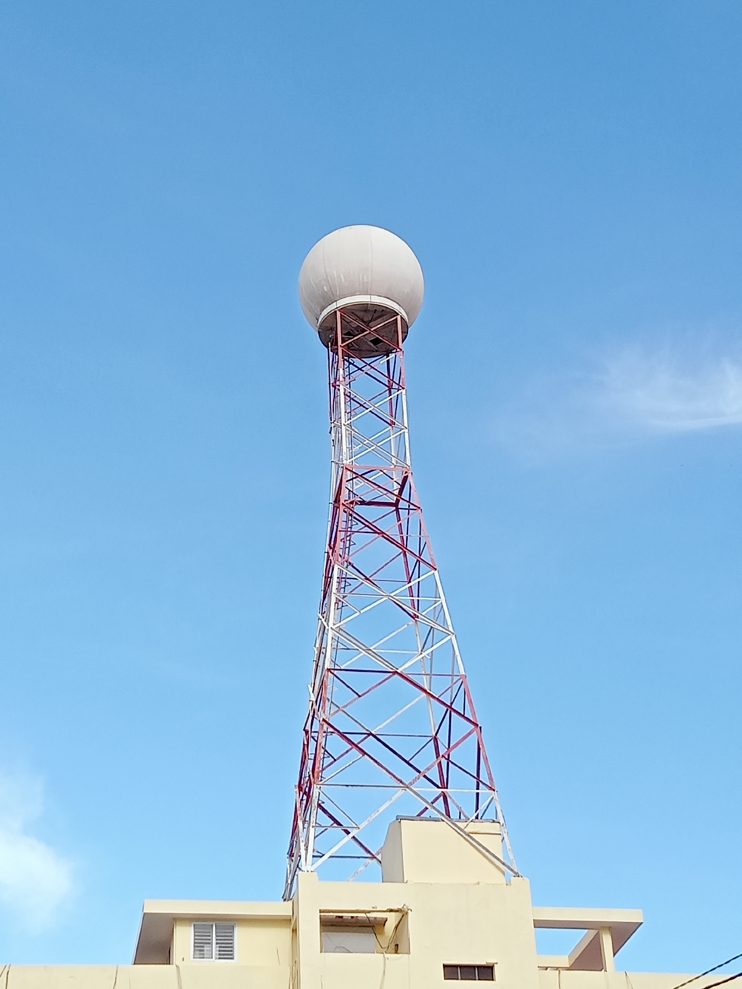 A tall, red and white lattice tower rises from the roof of a light yellow building. The tower is topped with a white radome. The sky behind the tower is clear and blue. The building has several windows, including one with white blinds. The image was taken in Nha Trang Ward, Vietnam.