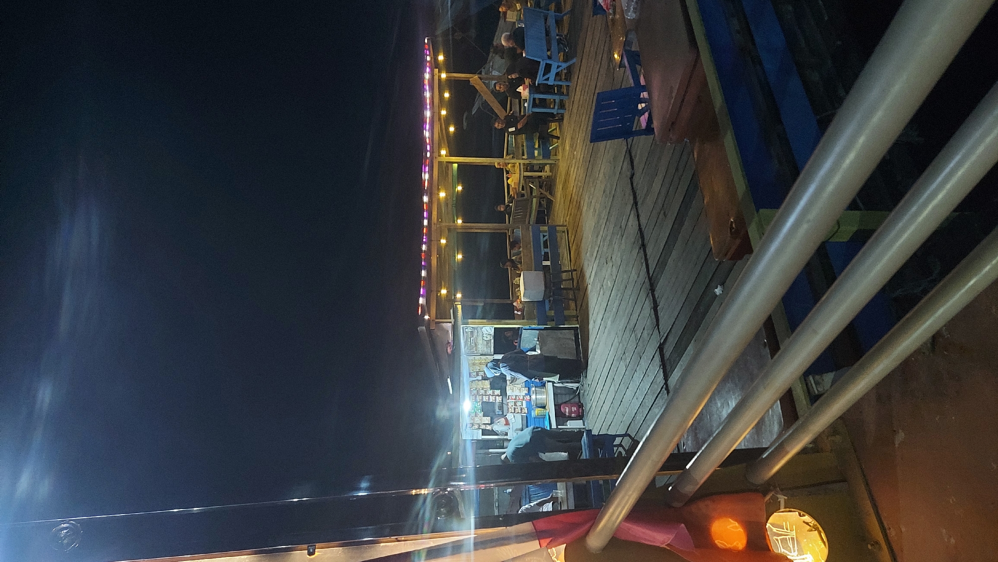 This is a nighttime photograph of an outdoor seating area at what appears to be a waterfront cafe or restaurant in Sambaliung, Indonesia. The scene is illuminated by string lights overhead and a bright light source to the left, casting long shadows on the wooden deck. In the foreground, a metal railing runs horizontally, with a partially visible red and white flag attached to its left.
Further back, a small food stall or counter is visible, with at least two individuals attending to it. One person in a blue hooded garment stands behind the counter, while another person in a lighter colored shirt appears to be interacting with them. Shelves behind the counter display various items. To the right of the stall, a covered seating area with blue benches and tables is set up. Several groups of people are seated at these tables, appearing to be dining or socializing. The overall atmosphere suggests a casual evening gathering. The wooden deck flooring extends throughout the visible area.