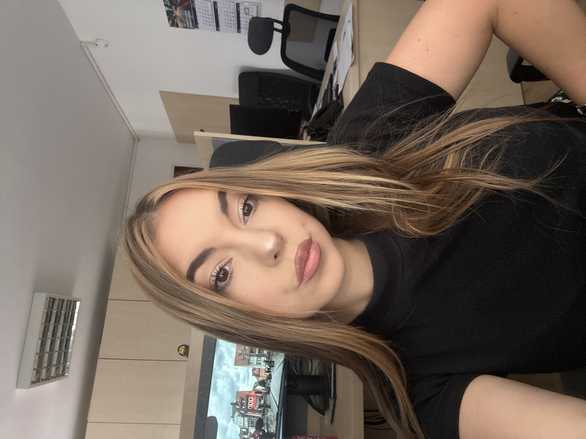 In a brightly lit office setting, a young woman with long, blonde-streaked brown hair is positioned in the foreground, looking directly at the camera. She is wearing a black, short-sleeved, high-neck ribbed top. Her makeup includes defined eyebrows, eyeliner, mascara, and pink lipstick. Behind her, a computer monitor displays an image of a European town with red-roofed buildings and a bridge. The desk she is seated at is visible, with papers and a phone. Further back, office furniture including two black office chairs and wooden cabinets are present. A fluorescent light fixture is mounted on the ceiling, and a calendar with images is on the wall. The overall impression is that of a casual selfie taken in an office environment, with elements of a typical workplace decor.