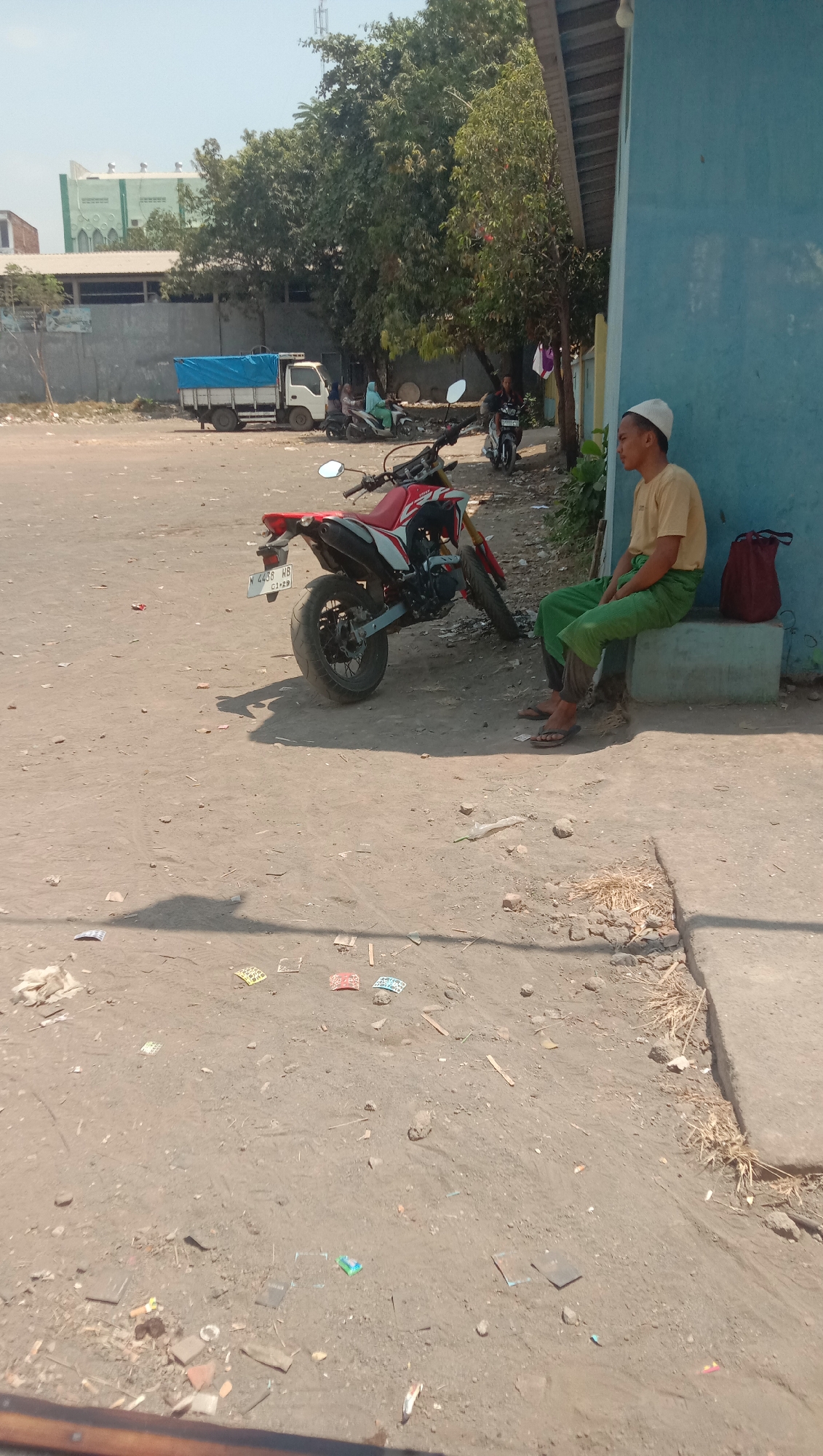 The image depicts a scene on a dusty, unpaved area in the city of Karanganyar, Indonesia. In the foreground, a red and white motorcycle is parked, with its license plate visible. To the right, a young man wearing a white skullcap, a yellow t-shirt, and green pants is sitting on a concrete ledge. He is looking to his left, away from the camera. A red backpack is placed on the ledge next to him.
In the background, a white truck with a blue tarp cover is parked. Several people are near the truck, some on motorcycles. A building with a green facade and white architectural elements, possibly a mosque, is visible further in the background. The environment appears to be an open lot with scattered debris on the ground, including paper scraps and other small objects. Trees with dense foliage are present in the mid-ground and background. The sky is clear and blue, indicating a sunny day.