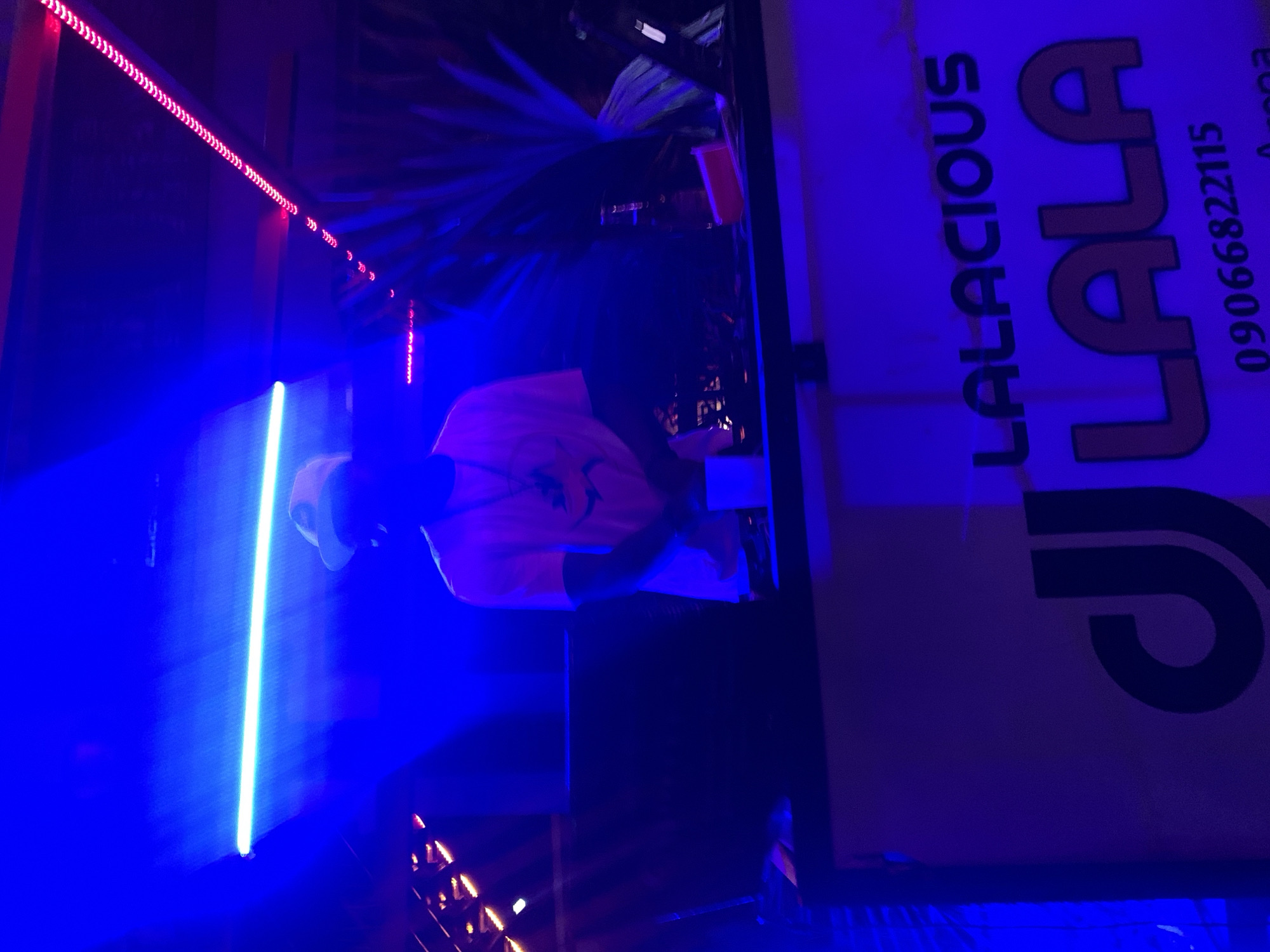 The image depicts a dimly lit indoor scene, likely a nightclub or event venue, bathed in vibrant blue and pink lighting. A person wearing a white cap and a light-colored t-shirt stands behind a DJ booth. The DJ booth has a large, stylized logo that reads "DJ LALA" in bold, outlined letters, with "LALACIOUS" printed above it. Below the logo, a phone number, "09066822115," is visible. The overall ambiance suggests a lively, possibly music-focused event taking place. The mention of Shendam, Nigeria, indicates the geographical location of this scene.