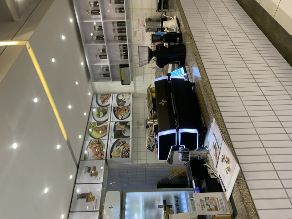 The image shows a counter in a cafe, located at coordinates -7.941 latitude, 112.608 longitude, which is in the city of Malang, Indonesia. The counter is made of light grey tiles and has a black and white espresso machine with the words "ALLEGRA MACCHINE ESPRESSO ITALIANE" on its front. There are several glass jars with coffee beans on a shelf above the counter. There are also several empty glass jars and a large black coffee grinder on the counter next to the espresso machine. To the left of the espresso machine are two large black cash registers and a sign that says "CASHLESS NOTICE". Behind the counter, on the wall, are multiple white framed pictures showcasing various types of food and coffee. The cafe has a white tiled ceiling with recessed lighting.