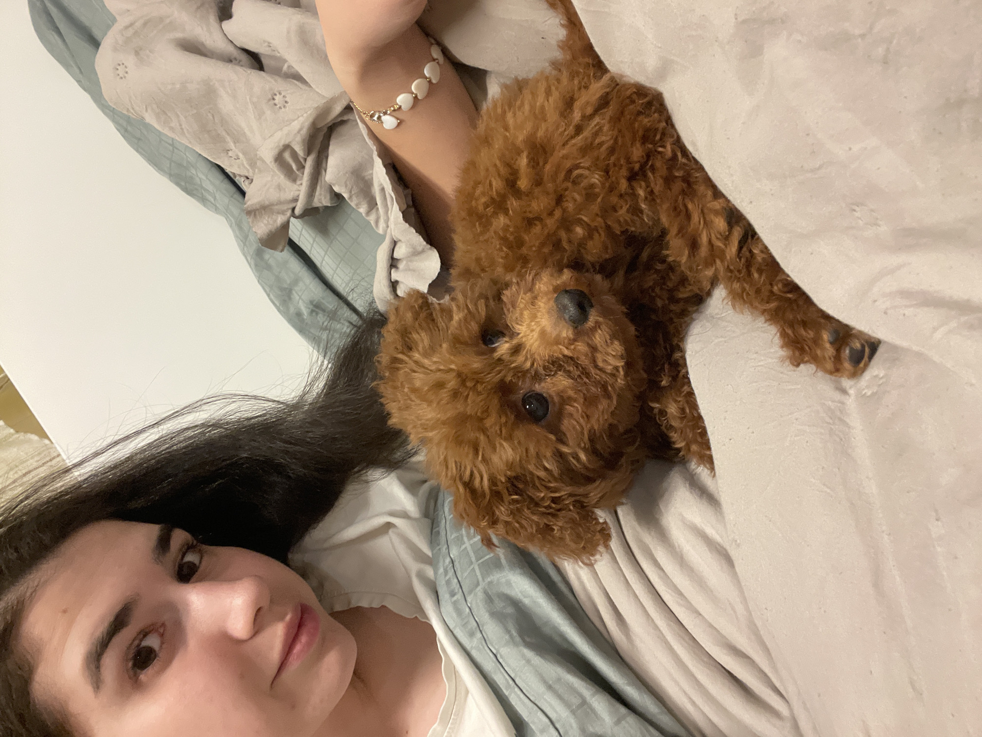 A woman with long, dark hair is lying on her back in a bed, with a small, brown poodle lying on her torso. She is wearing a white shirt and a bracelet with heart-shaped beads on her left wrist. The bed is covered with gray and white linens. The location cannot be determined as it is not visible in the image.