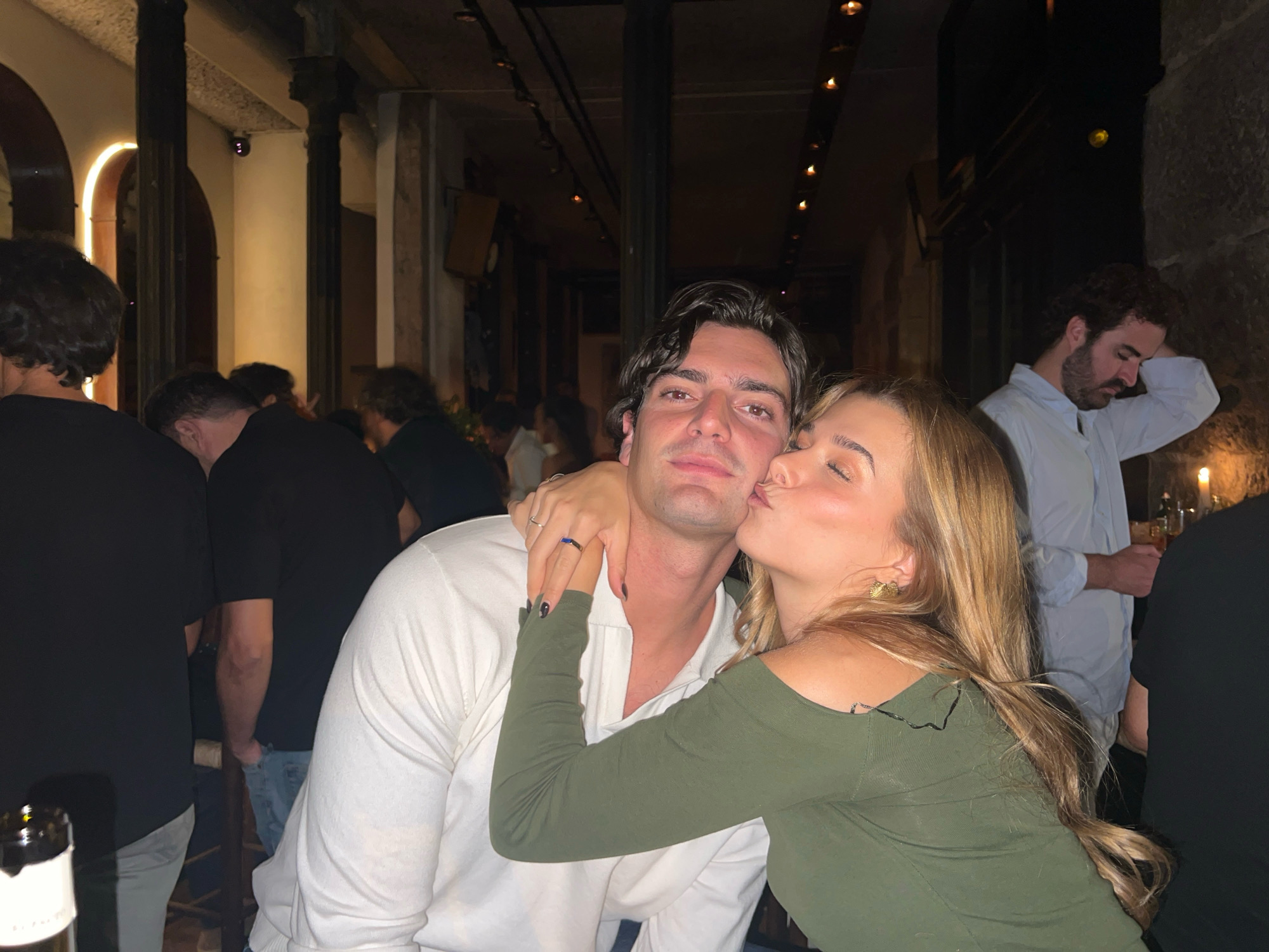 The image shows a couple kissing indoors. The man is wearing a white shirt, and the woman is wearing a green top. They are in the foreground, with other people in the background. The environment appears to be a restaurant or bar with dim lighting, high ceilings, and visible architectural elements such as pillars and arches. The presence of decorative lighting fixtures further suggests an indoor dining or entertainment venue. The scene is likely in Madrid, Spain, given the context provided, but the image itself does not contain explicit visual cues to confirm this specific location. The focus is on the intimate moment between the couple, with the background blurred and less detailed.