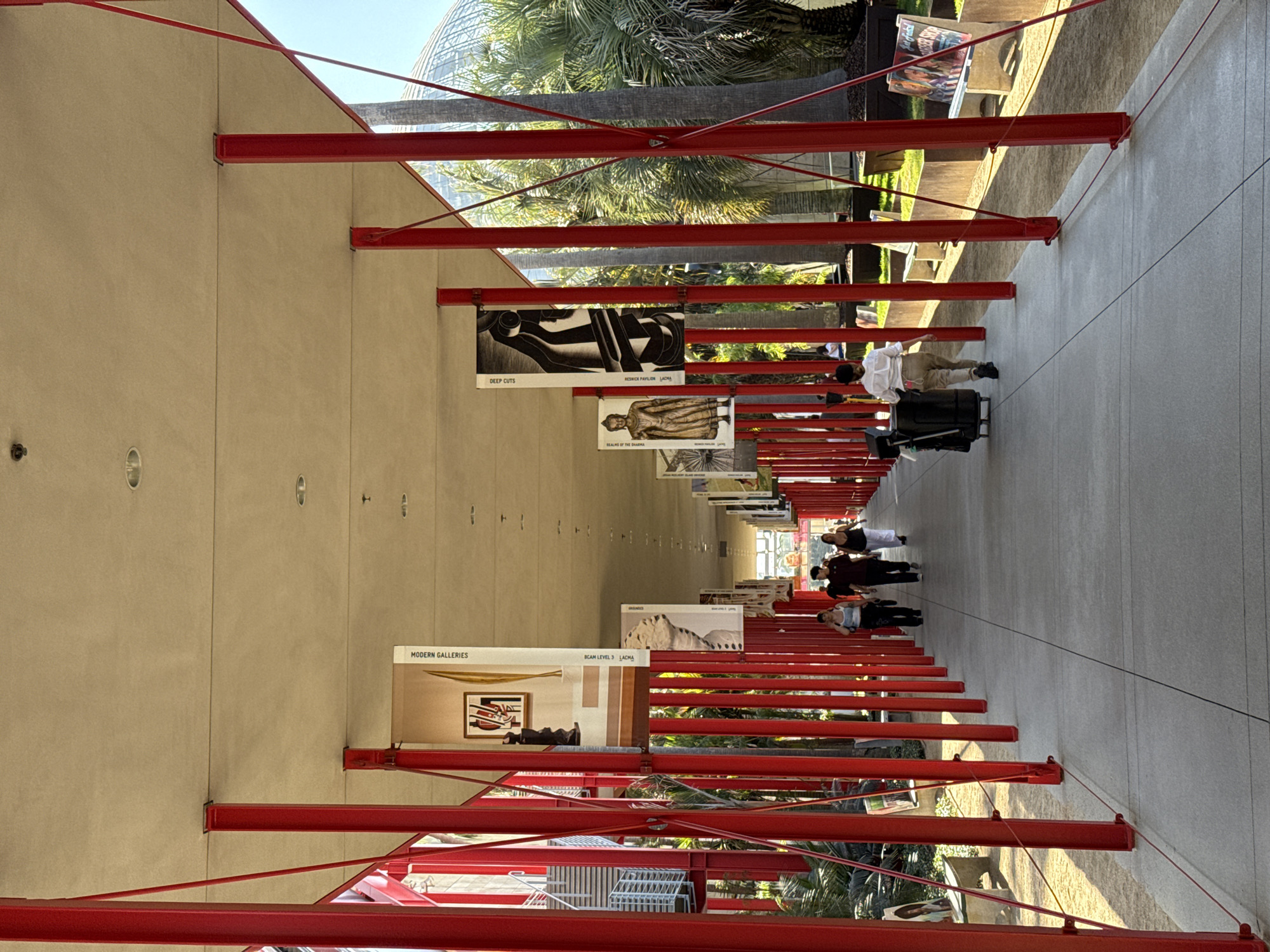 The image depicts a covered walkway with a row of vibrant red support posts on either side. The walkway itself is paved and appears to be an outdoor public space. Several people are walking along the walkway, some individually and some in small groups. In the background, palm trees and part of a domed structure are visible, suggesting a location with a warm climate. Hanging banners featuring artwork and text are displayed along the walkway, indicating a cultural or artistic venue. The lighting suggests it is daytime, with sunlight casting shadows.
This scene is consistent with the architecture and outdoor spaces found at the Getty Center in Los Angeles, California. The distinctive red columns and the artwork displayed are characteristic of the Getty's design and exhibitions. The presence of palm trees and the architectural style further support this identification.