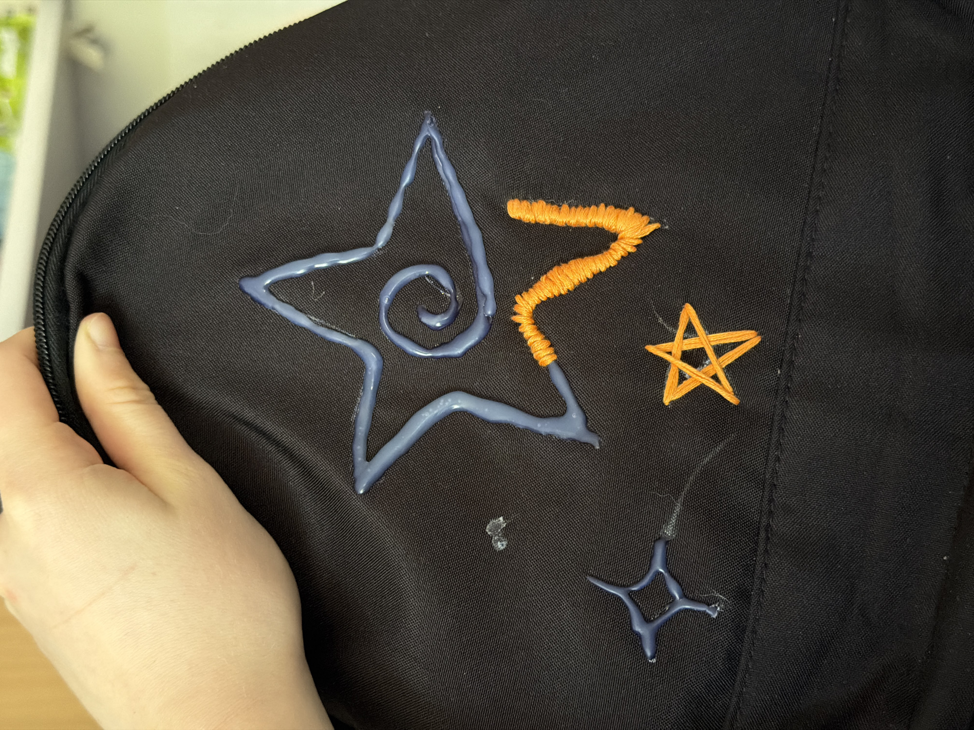 A close-up shot shows a black backpack or bag with stitched star designs. One large star is outlined in a shiny, light blue material, with a spiral design in the center. To the right of this, an orange zigzag shape is stitched. Further to the right and below are two smaller stars, one orange and one light blue, also created with a shiny material. A light-skinned human hand holds part of the bag on the left side of the frame. The background is indistinct, but a wooden surface is visible below the bag. The location cannot be determined from the image.