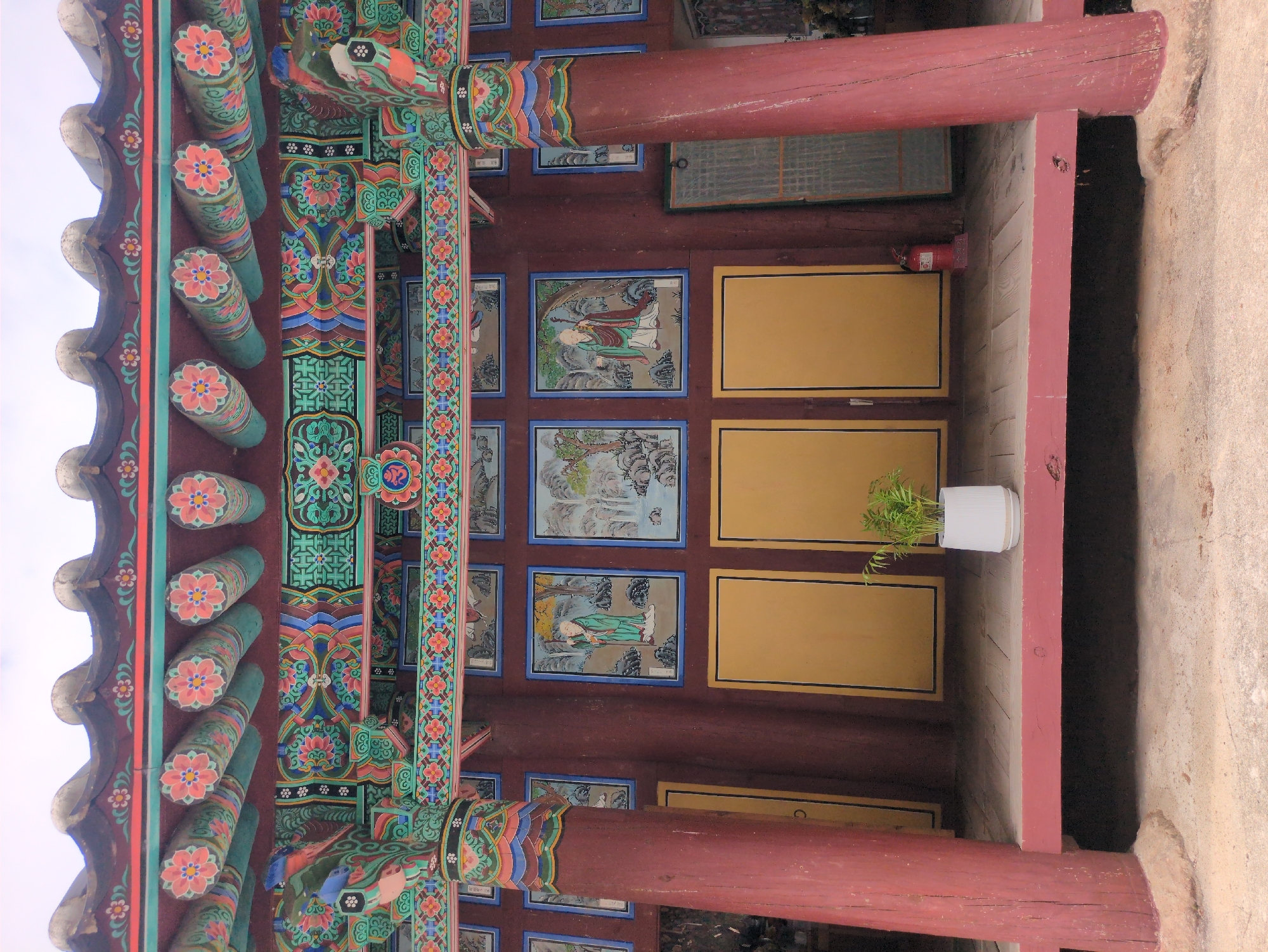 The image shows an ornate traditional Korean building, possibly a temple or palace, located in Gongju-si, South Korea. The structure features elaborately decorated eaves with colorful floral and geometric patterns on the bracketing system (Dancheong). Red wooden pillars support the roof. The interior reveals a series of framed paintings of figures and landscapes, interspersed with yellow rectangular panels. A small potted plant sits in front of a pair of yellow doors, and a red fire extinguisher is mounted on the wall nearby.