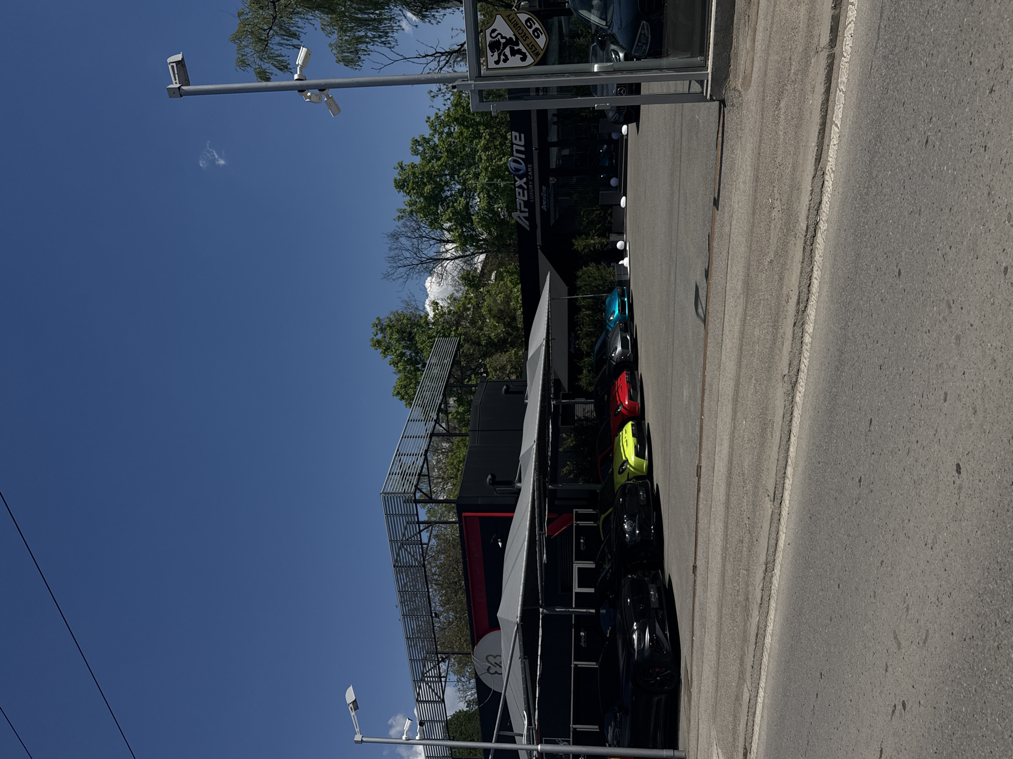 The image depicts a street scene with an "Apex One Luxury Cars" dealership, presumably located in Sofia, Bulgaria. The dealership features a modern, multi-level structure with a partially covered rooftop area. A row of cars, including a black, yellow, red, and blue vehicle, are visible next to the dealership along the street. The sky is clear with a few clouds. Streetlights are also visible.