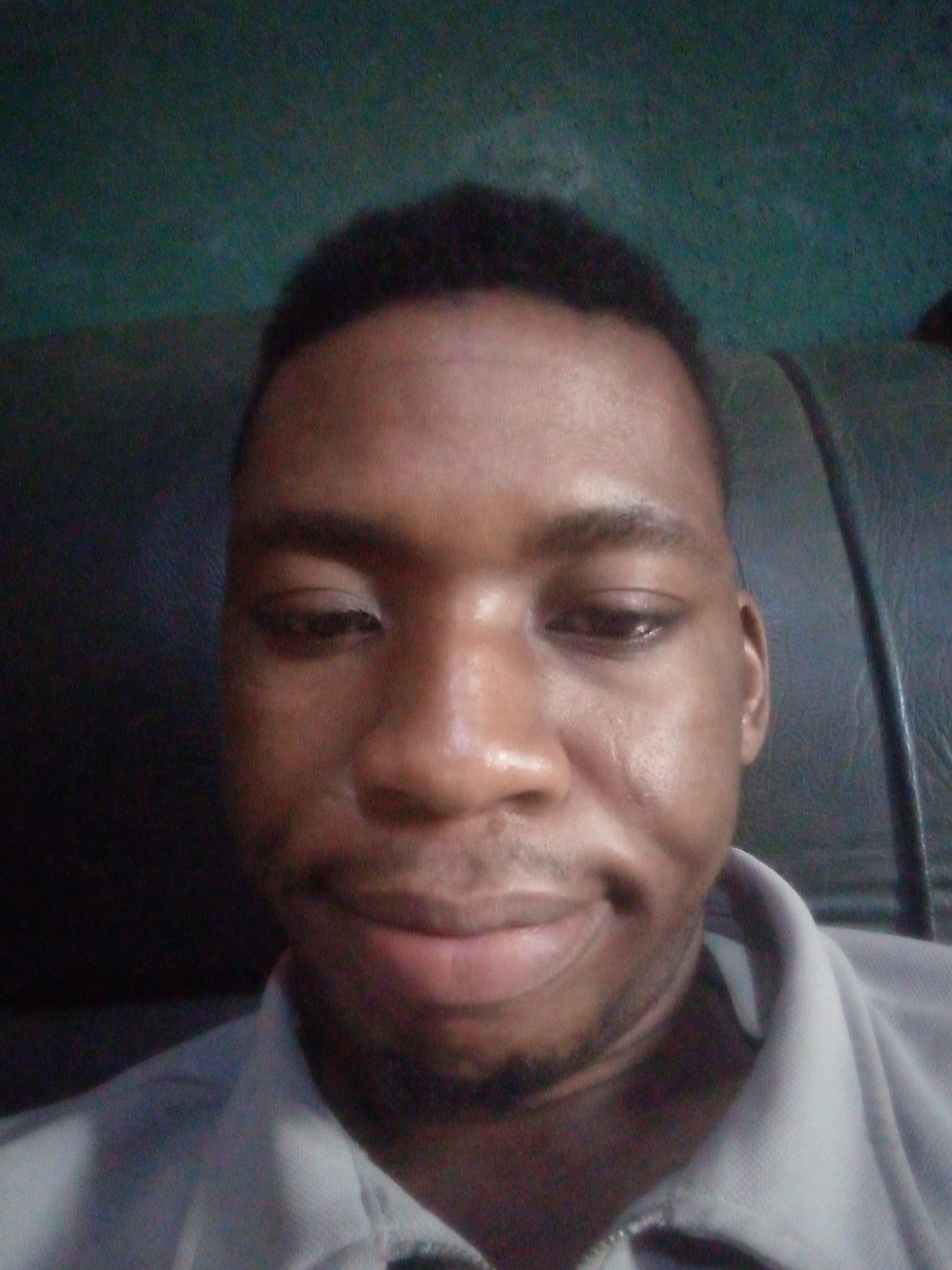 A close-up selfie of a dark-skinned man with short hair. He is wearing a light-colored shirt. The background is a blurry dark green wall and what appears to be a dark colored sofa. The location is unknown.