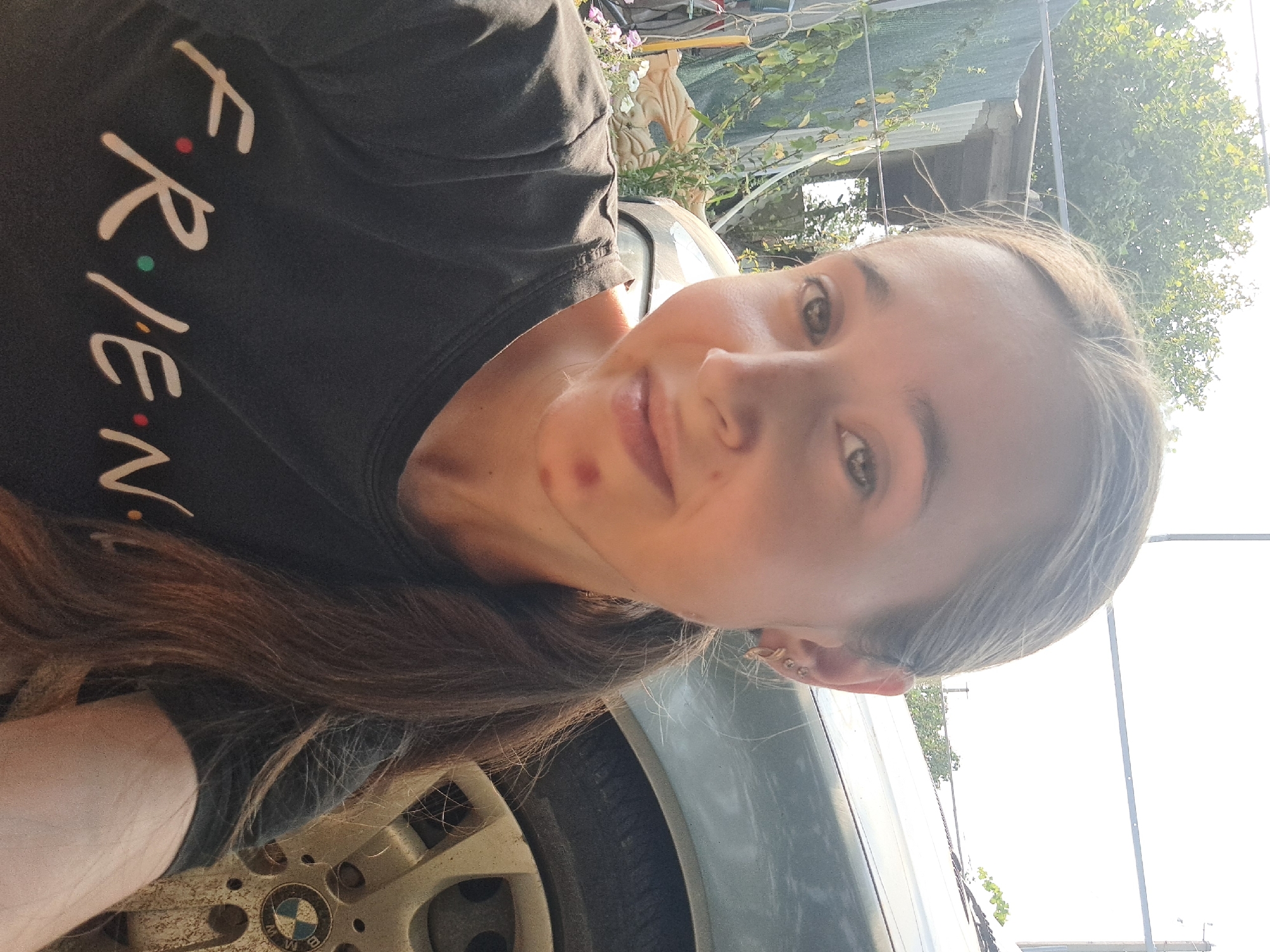 A young woman is captured in a selfie, her face turned slightly towards the camera. She has fair skin, light brown hair pulled back into a ponytail, and dark eyes. A small red blemish is visible on her chin. She is wearing a dark t-shirt with the word "FRIENDS" printed on it in white letters, with colorful dots representing the 'i's and the dot after 'E'. Behind her, a portion of a car, possibly a BMW given the emblem visible on the wheel, is present. The background suggests an outdoor setting, with greenery and what appears to be a fence or corrugated metal structure. The lighting indicates it is daytime, with bright sunlight casting shadows. The location is identified as Vypasne, Ukraine.