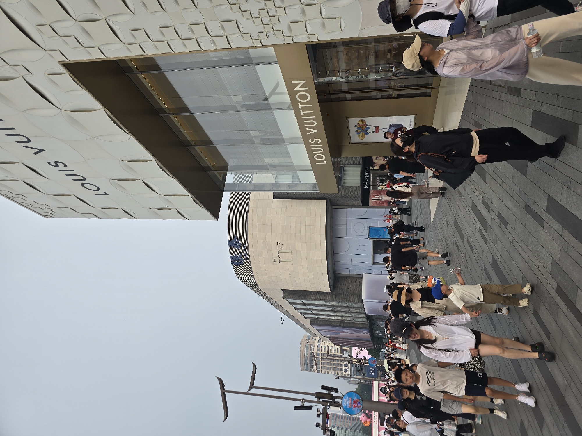 A street-level, eye-level, full shot captures a bustling pedestrian area in Shangcheng District, China, under an overcast sky. The foreground is dominated by a wide, patterned walkway with a dark grey and light grey tile design. To the right, the imposing facade of a Louis Vuitton store stands out, featuring a beige textured exterior with the store's name prominently displayed in gold lettering above a large glass storefront. To the left, other commercial buildings line the street, including a large structure with "in 77" branding and a curved, beige facade. Further in the distance, taller city buildings are visible.
A diverse crowd of people walks along the patterned pathway. In the immediate foreground on the left, a group of individuals are walking together: a young boy in a white t-shirt and blue cap, holding a woman's hand; the woman, wearing a white jacket and black shorts; and a teenage boy in a grey t-shirt and dark shorts. To their right, a woman in a black jacket and black pants walks by, carrying a large black tote bag. Further to the right, a person in a light purple jacket and beige pants walks with a water bottle. Behind them, numerous other pedestrians are seen in various directions, some carrying shopping bags. Streetlights with curved tops and surveillance cameras are mounted on poles along the left side of the walkway. Promotional posters are visible on some of the building facades. The scene depicts a commercial district during daylight hours.