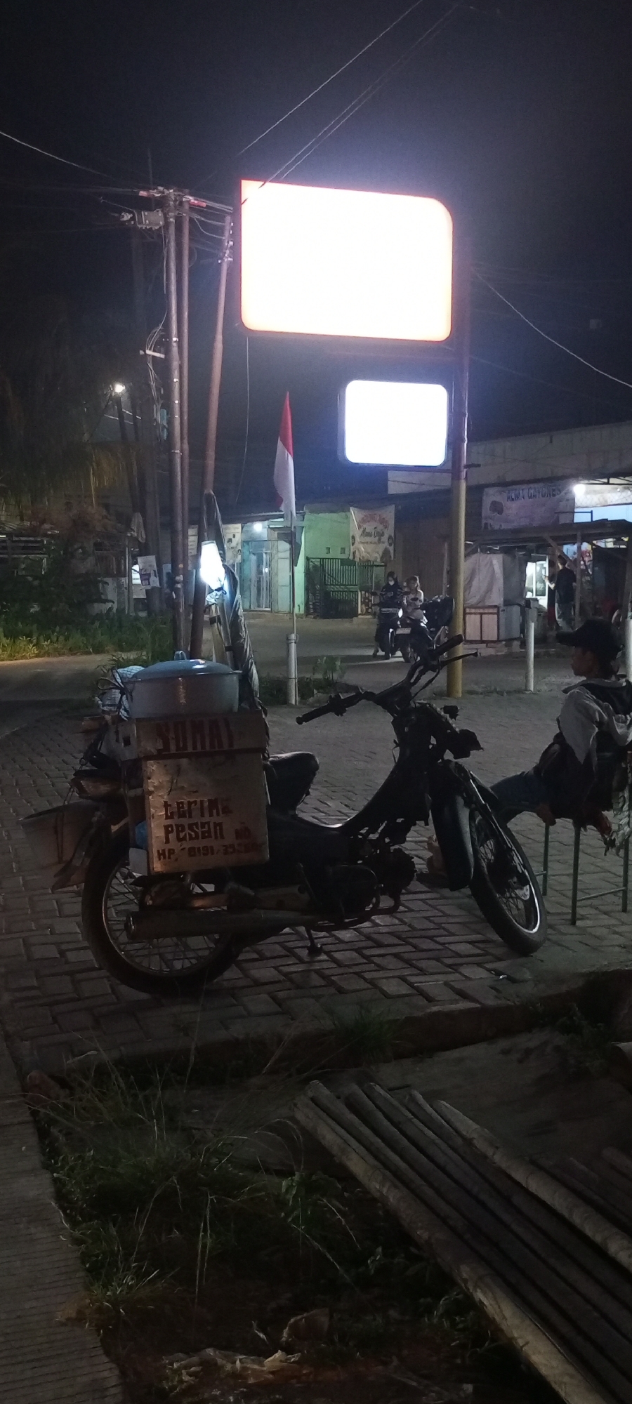 A nighttime exterior shot captures a street scene in Setu, Indonesia. A parked motorcycle with a large metal container attached to its rear is the central subject. The container is adorned with text, indicating "SUMAY" and "TERIMA PESAN" (meaning "accept orders"), along with a phone number "HP: 0191 / 3528" which is partially obscured. To the right of the motorcycle, a person is seated on a metal chair. In the background, illuminated by streetlights and overhead signage, are various buildings and a street with a few other motorcycles and people visible. An Indonesian flag is prominently displayed, waving from a pole. The ground is paved with interlocking bricks.