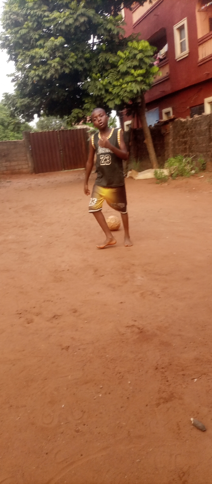 The image displays a young male individual standing on a reddish-brown dirt surface in Ogidi, Nigeria. The boy, who appears to be of adolescent age, is barefoot and wears a dark sleeveless jersey featuring the number "23" and text, possibly "LAKERS," with accompanying dark and yellow/orange patterned shorts. He is positioned with his right foot adjacent to a worn, light-colored soccer ball resting on the ground. His gaze is directed towards the camera. The background on the left features a low concrete wall topped with a section of reddish-brown corrugated metal fencing. Dominating the upper left and center background is a large tree with dense green foliage. To the right, a multi-story building, painted reddish-brown, is visible, featuring white-framed windows and several balconies. The overall environment is an unpaved outdoor area with residential structures and natural vegetation.