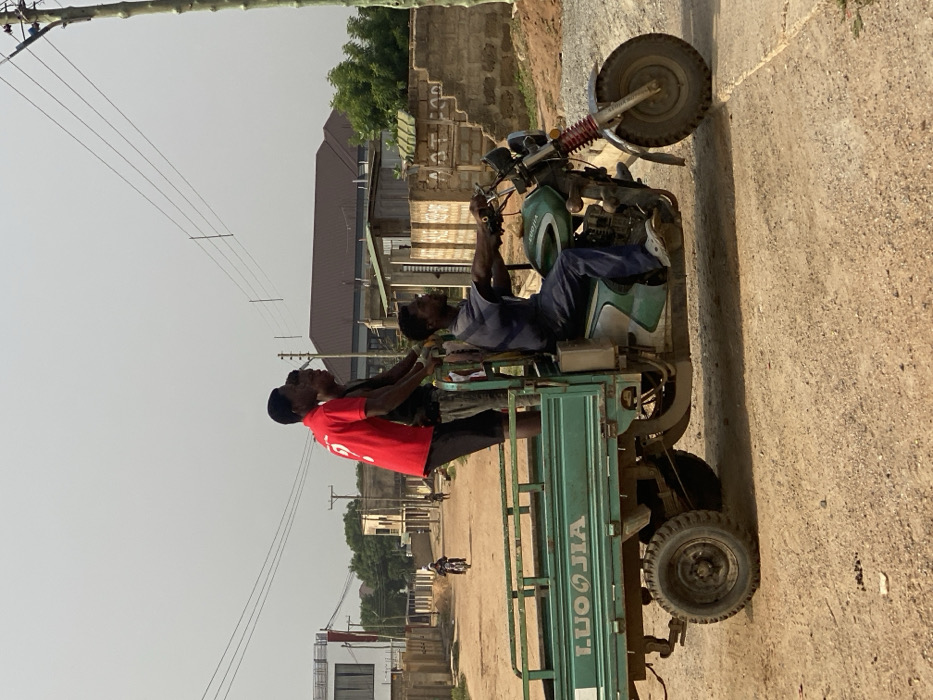 The image depicts a green three-wheeled motorized cargo vehicle, a "LUOJIA" brand, traversing an unpaved, dusty dirt road in Joma, Ghana. Four individuals are on board. A male driver, wearing a blue shirt and pants with white Crocs, is seated on the motorcycle portion, operating the handlebars. In the attached cargo bed, three male passengers are present. Two of them are standing upright; one wears a red t-shirt with a white '9' on the back and black shorts, while the other wears a dark t-shirt and dark shorts. A third passenger is partially visible, positioned lower in the cargo bed behind the other two.
The environment indicates a residential or semi-urban area. In the background to the left, a multi-story white building with dark windows is visible, along with other concrete block structures and utility poles supporting overhead wires. Further down the road, another motorcycle and individuals are discernible. To the right, a partially damaged concrete block wall stands alongside another utility pole. The sky is a light, hazy gray, suggesting an outdoor setting during daylight. The ground features dry, light-brown soil, with a visible concrete edge on the immediate right.