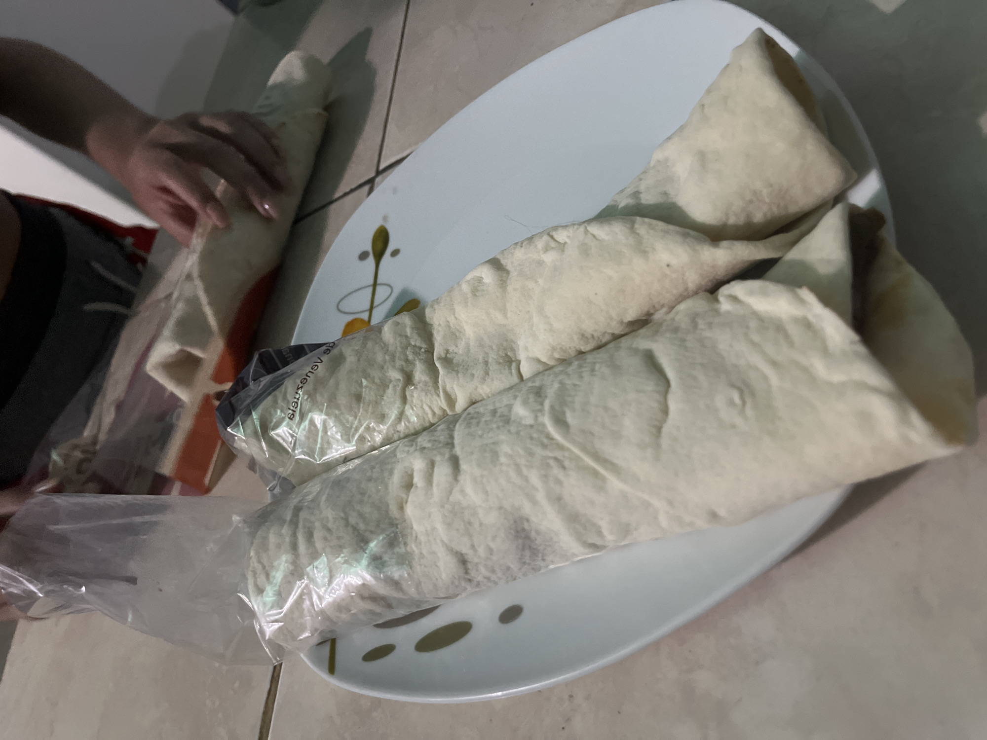 The image captures an indoor scene in Juan Griego, Venezuela, likely a kitchen or dining area, where food is being prepared or unwrapped. The primary subjects are a person and several food items resembling burritos or wraps.
In the foreground and center, a white plate holds three large, rolled food items, possibly burritos or shawarmas, made with a light-colored flatbread. Two of these are partially encased in clear plastic packaging. The plate itself is white with subtle decorative elements in green/yellow and black dots. To the left, a person's hands are visible, in the process of unwrapping or preparing another similar food item, which is also rolled and appears to be in clear plastic. The background shows light-colored tiled flooring and a wall.
The activity centers around handling and preparing these rolled food items. The lighting is somewhat dim, casting shadows, suggesting it might be evening or an indoor setting with artificial light. On the plastic packaging, reversed text is visible, which, when flipped, appears to read "ensuenos" or similar.