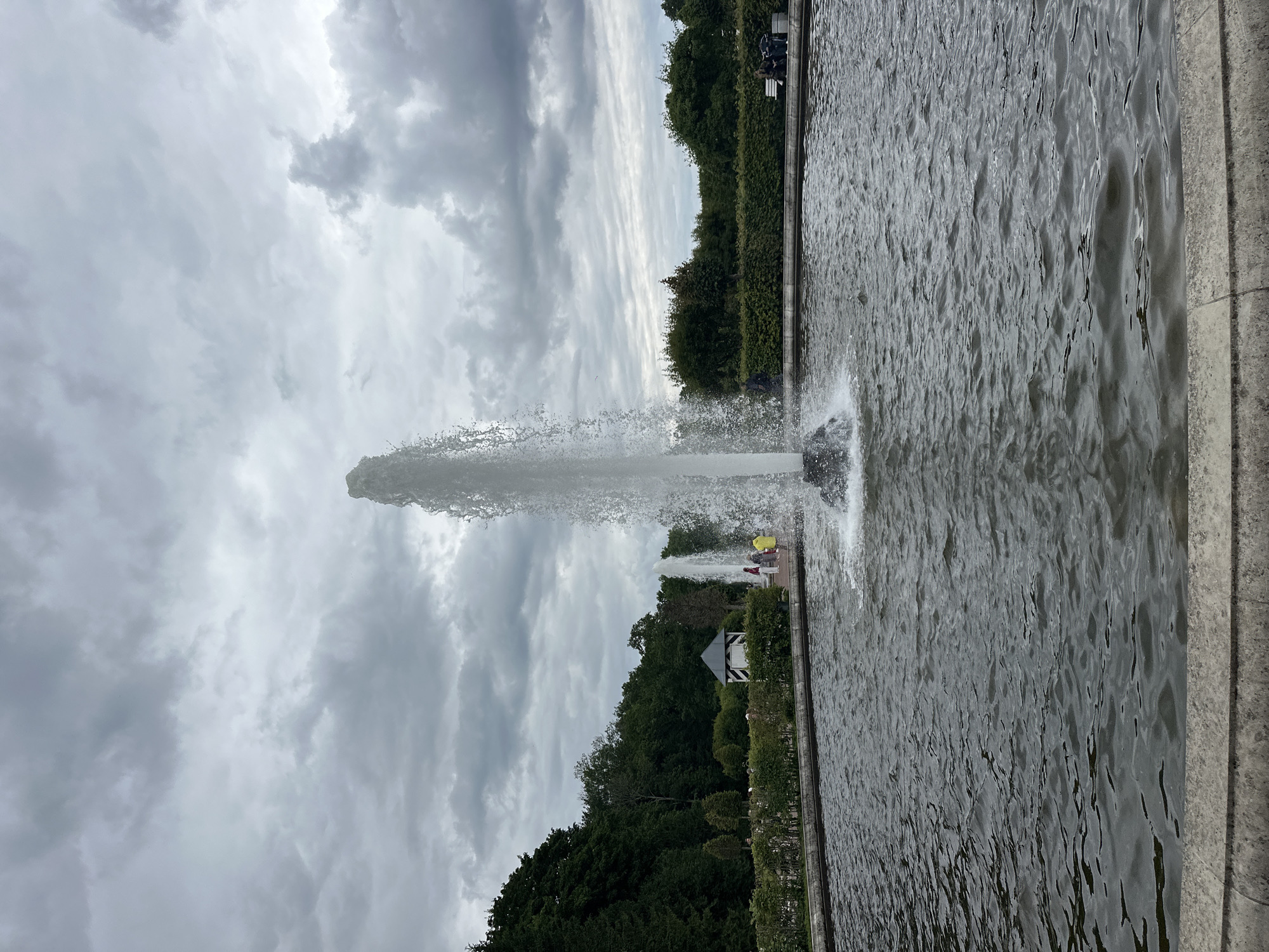 The image shows a large fountain in a pond at Peterhof, Russia. The fountain is shooting a tall stream of water into the air. The pond is surrounded by manicured hedges and trees. In the background, there are several people walking and sitting. The sky is overcast and cloudy.