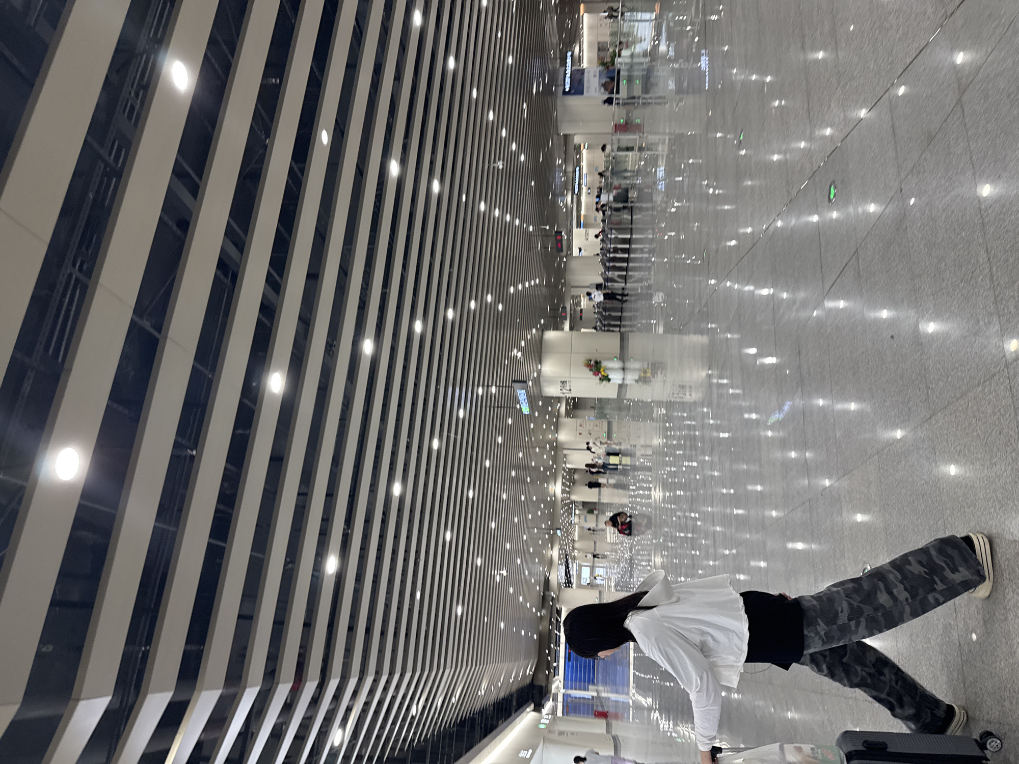 A young woman with long black hair is walking with a suitcase in a modern, well-lit airport terminal, likely located in Baiyun District, China. She is wearing a white hooded jacket over a black top and camouflage pants. The floor of the terminal is highly reflective, creating numerous light spots from overhead lighting and the ceiling's slatted design. In the background, other travelers are visible, some walking and others seated. Security gates and signage are also present in the distance. The overall environment suggests a busy transportation hub.