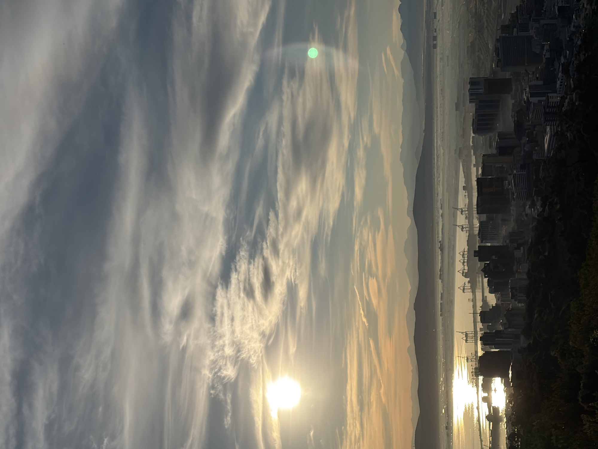 The image depicts a panoramic view of Cape Town, South Africa, at sunrise or sunset. The foreground shows a densely packed urban landscape with numerous buildings of varying heights, some appearing to be commercial or residential. A prominent port with large cranes is visible in the mid-ground, situated along a body of water that reflects the warm hues of the sky. In the background, a range of mountains forms a natural backdrop to the city. The sky is filled with dynamic cloud formations, illuminated by the low-lying sun, which casts a golden glow over the scene. A peculiar green orb-like anomaly is present in the sky to the right of the sun. The overall atmosphere is one of urban development juxtaposed with natural landscape, bathed in dramatic lighting.