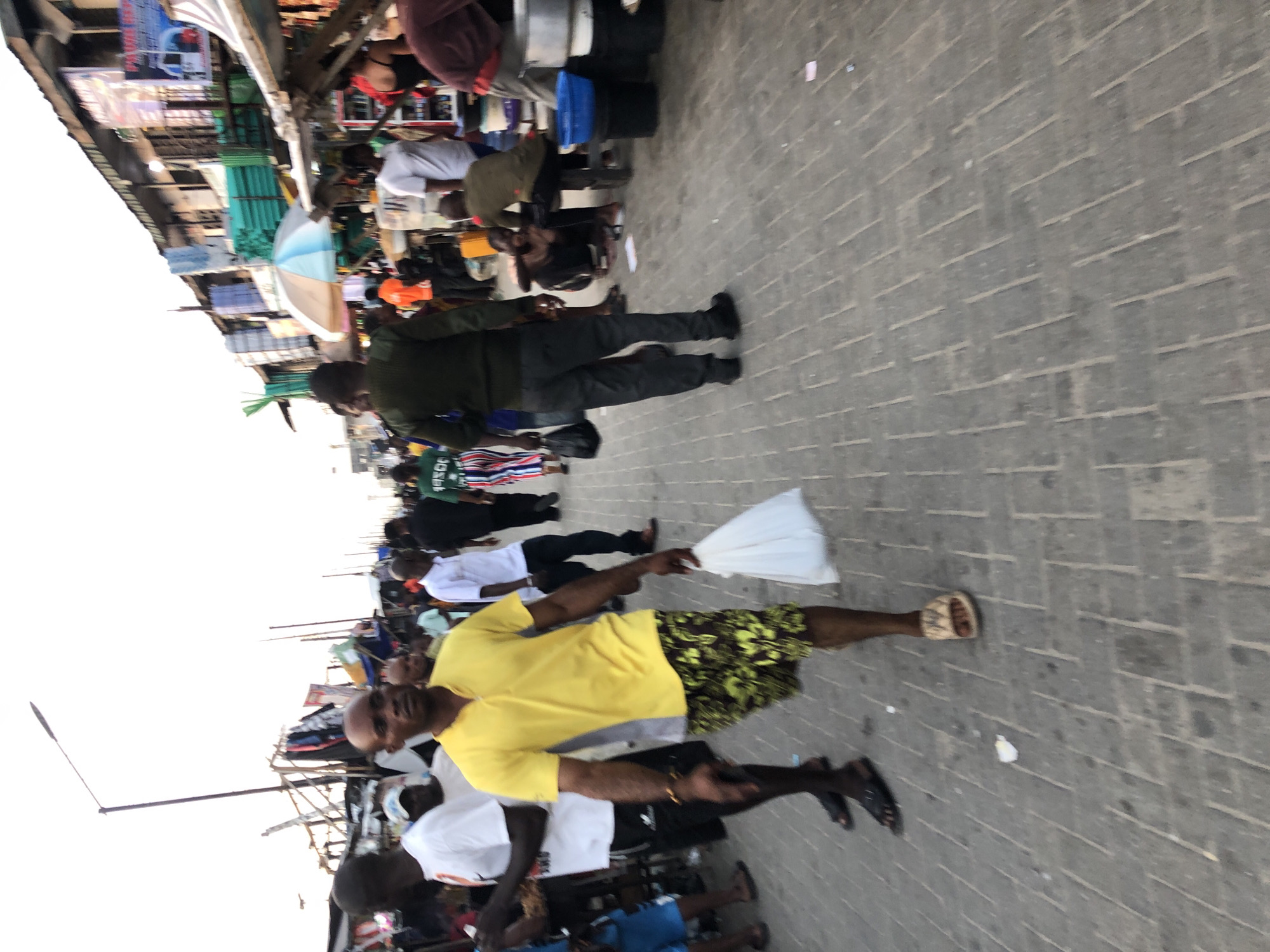 The image depicts a crowded street scene in Moba, Nigeria. The location appears to be a market area with stalls lining the left side of the frame. A variety of individuals are present, with most of them walking along the paved street. A man in a yellow shirt and floral shorts holds a white plastic bag. Street lights are also visible on the left.