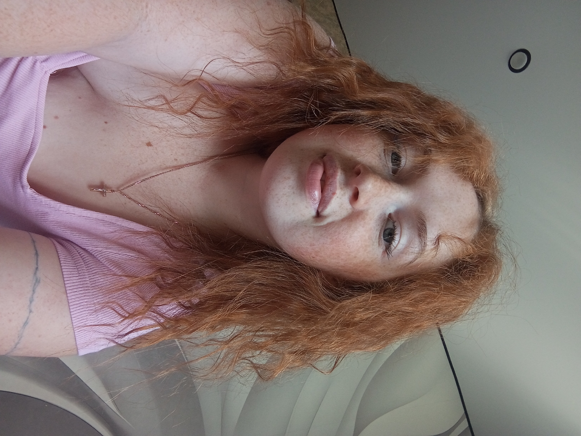 A young, fair-skinned woman with red curly hair and freckles poses for a selfie. She wears a light pink top and a delicate necklace with a cross pendant. The background is a plain wall, possibly indoors. The photo appears to be taken in a casual setting. There is no verifiable information about the location in Yurgamysh, Russia, or any actions, interactions. The image focuses on the woman's appearance and is seemingly devoid of elements indicating a specific geographical location.