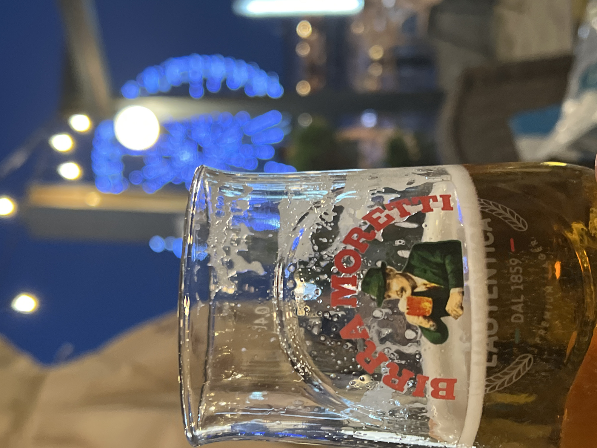 A close-up, low-angle shot focuses on a partially filled glass of beer, with a foamy head, resting on a surface. The glass bears the "Birra Moretti" logo, featuring a man in a green hat and suit holding a mug of beer. Text on the glass reads "AUTENTICA" and "DAL 1859" along with "PREMIUM LAGER". The background is blurred and shows a night scene with bright blue and yellow lights, suggesting an outdoor setting, possibly a boardwalk or entertainment area. The lights appear to be strung along poles and structures. The environment suggests a public space, potentially a restaurant or bar area, at dusk or nighttime. The bokeh effect of the lights implies a shallow depth of field. There are no visible people interacting with the glass. The overall impression is a casual, evening setting, likely in a city or resort area. The image does not contain any specific landmarks that would definitively place it in Constanța, Romania.
