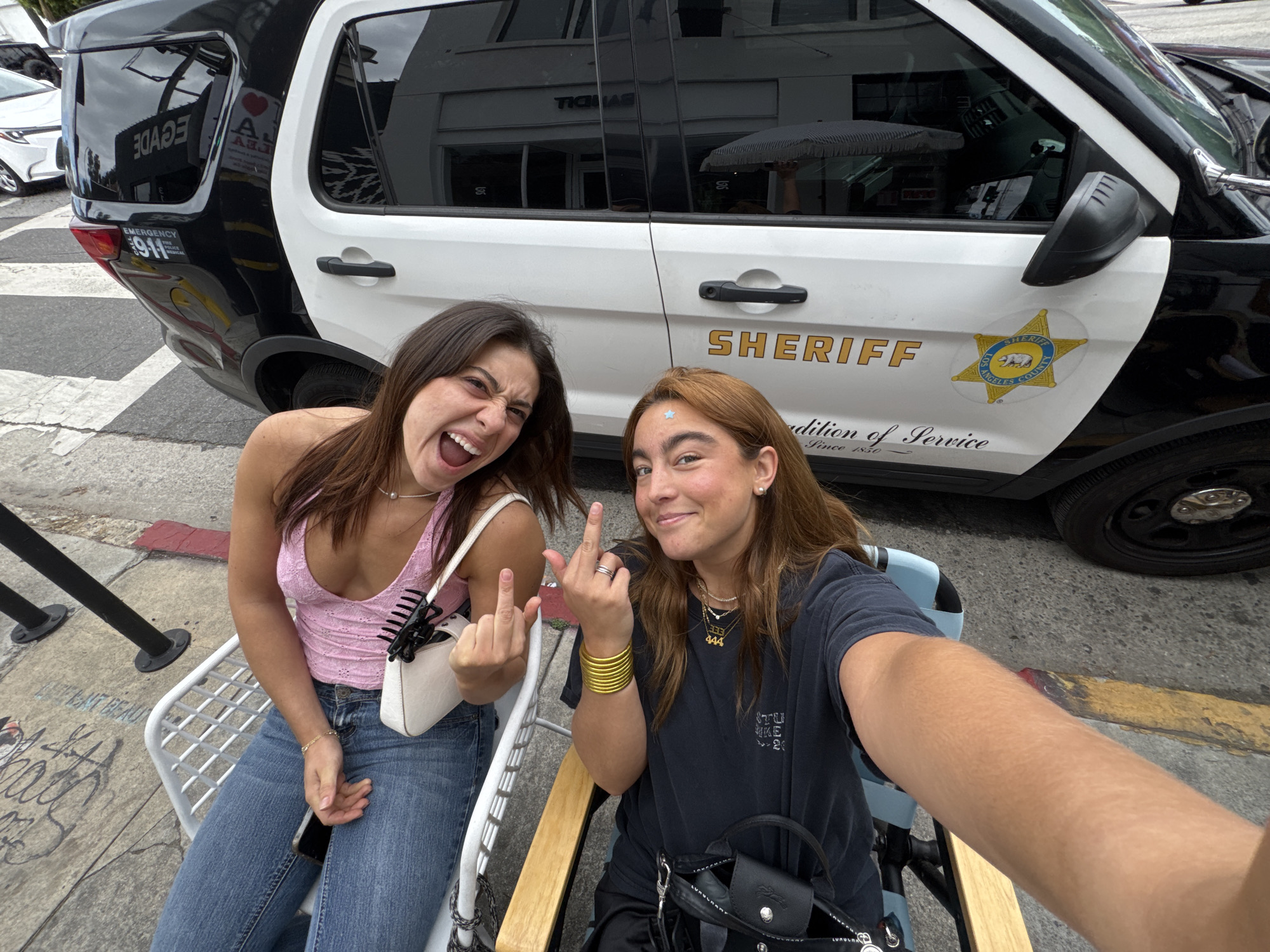 Two young women are seated in chairs, flipping off the camera while positioned in front of a Los Angeles County Sheriff's patrol vehicle. The car, marked "SHERIFF" and adorned with the Los Angeles County seal, indicates a location within Los Angeles County, United States of America. The presence of street-level seating and pedestrians passing by suggests an outdoor public space. Graffiti is visible on the sidewalk nearby.