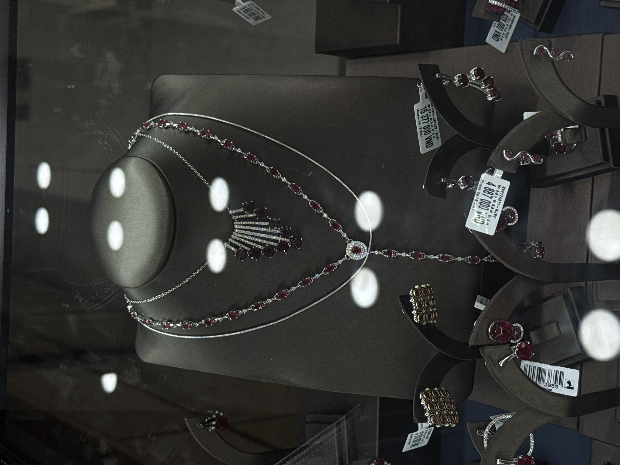 A jewelry store display case in Nha Trang Ward, Vietnam, showcases necklaces and earrings. A dark gray, neck-shaped stand in the center displays a long silver necklace with red ruby-like stones. Above this piece, a more elaborate necklace featuring a fan-shaped central ornament with matching red stones is visible.
To the right, various earring sets are arranged on dark brown stands. The earrings are also silver with red stones. Price tags, displaying amounts in Vietnamese Dong (VND), are attached to many of the pieces. The display case is illuminated with bright lights, creating reflections on the glass surface.
