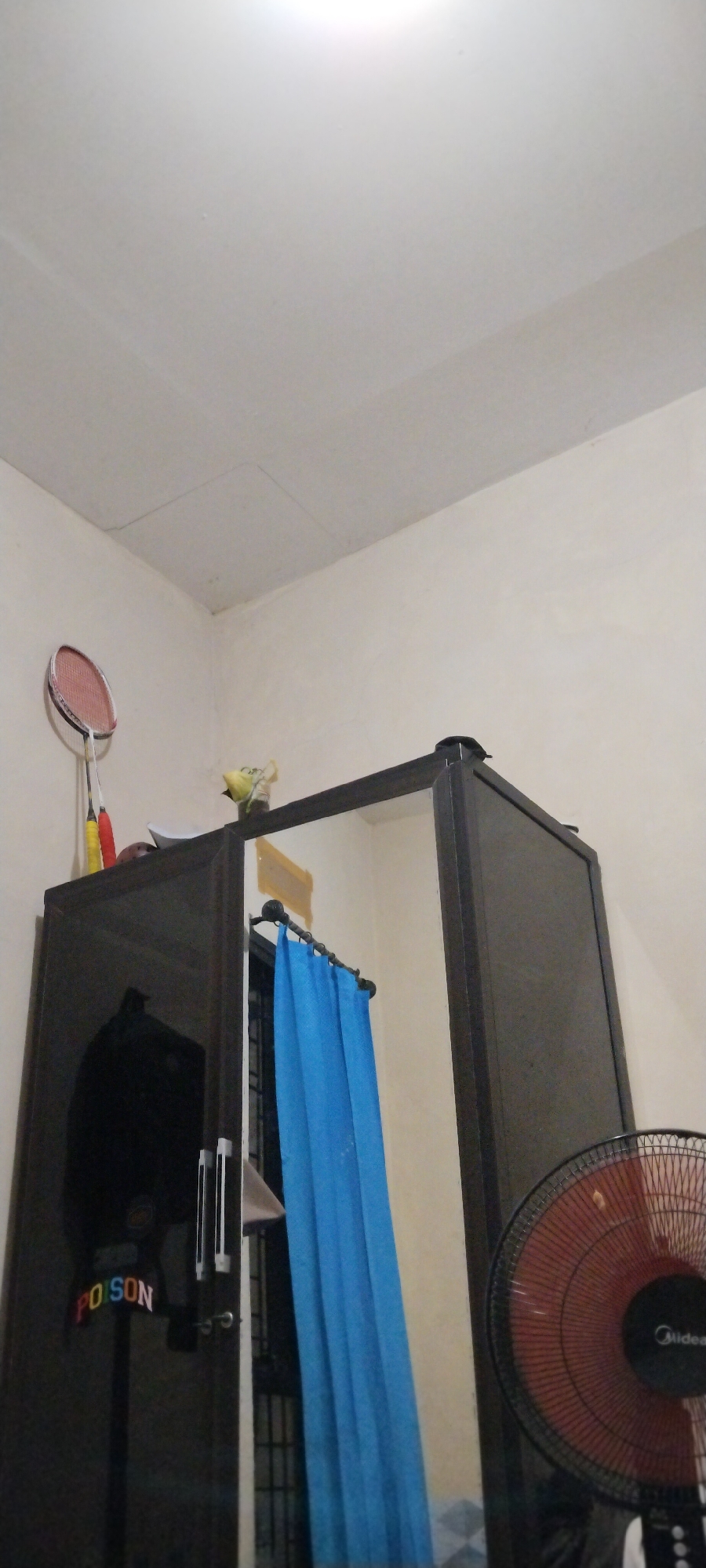 The image depicts an interior scene, likely a bedroom or storage area, with a focus on a dark brown wardrobe. A badminton racket with a red frame and white strings is leaning against the wall on the left side of the wardrobe, with two shuttlecocks also visible. On top of the wardrobe, a small, clear glass vase contains a yellow object that appears to be a fake flower.
The wardrobe itself has a mirrored door on the left, reflecting a portion of the room and showing two handles. A colorful sign with the word "POISON" is attached to the mirrored door. To the right of the mirrored door, there is an open section of the wardrobe, revealing a blue curtain hanging on a rod. The right side of the wardrobe has a solid, dark brown door. In the foreground on the right side of the image, a black oscillating fan with a red grill is visible. The walls are painted a light cream color, and the ceiling is white. There is a square-shaped access panel visible on the ceiling.
The specific location in Duman, Indonesia, cannot be determined from the visual information provided in the image.