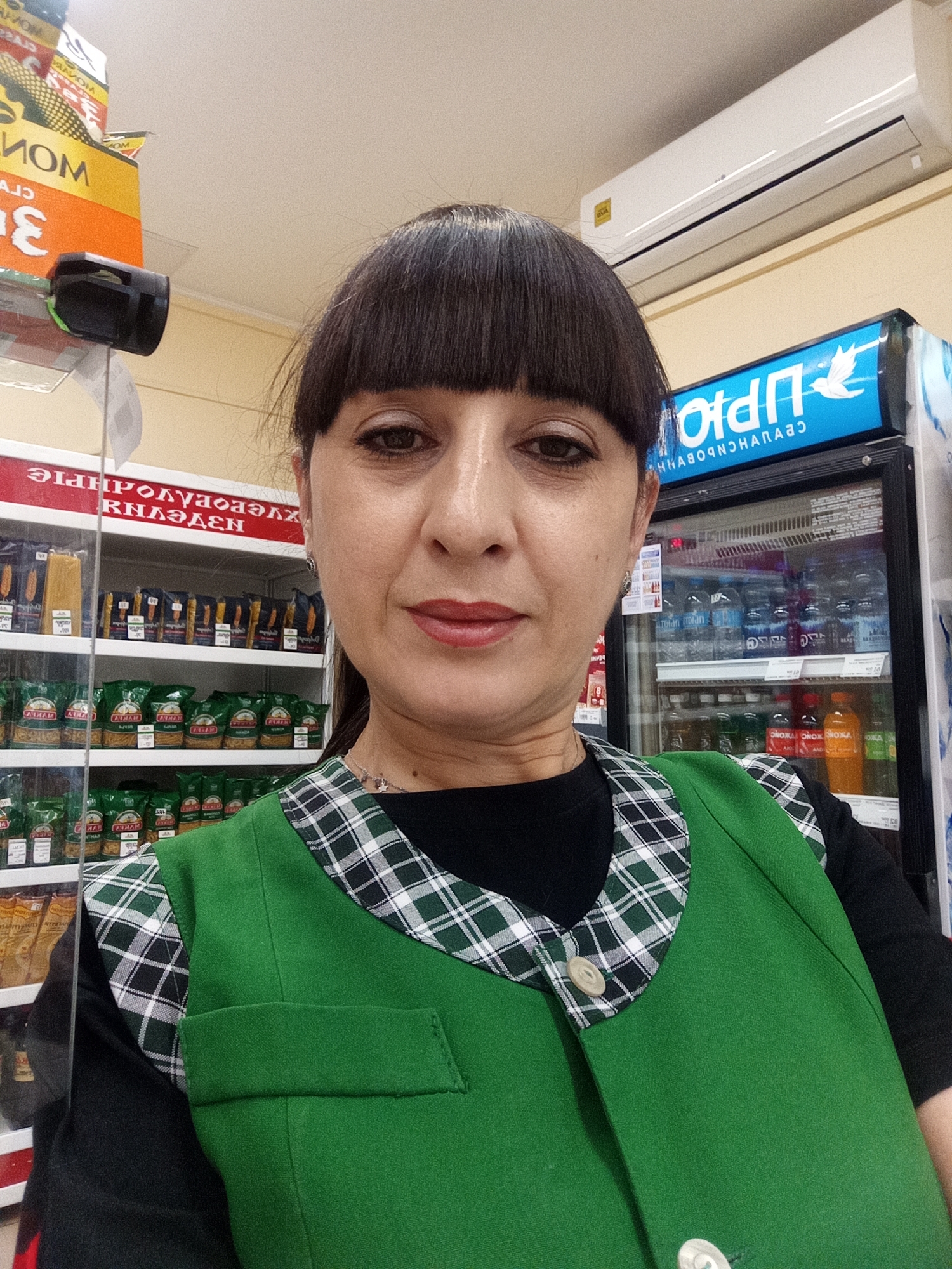 A woman with dark hair and bangs, wearing a green vest over a black shirt, stands in what appears to be a retail store. She is looking towards the camera and smiling. Behind her, shelves are stocked with packaged food items, including pasta. To her right, a refrigerated display case contains bottles of beverages. An air conditioning unit is mounted on the wall above the display case. The overall scene suggests a casual, everyday setting within a shop. The image is likely taken in Belogorsk, Russia, given the presence of Cyrillic text on the shelving and the overall context.