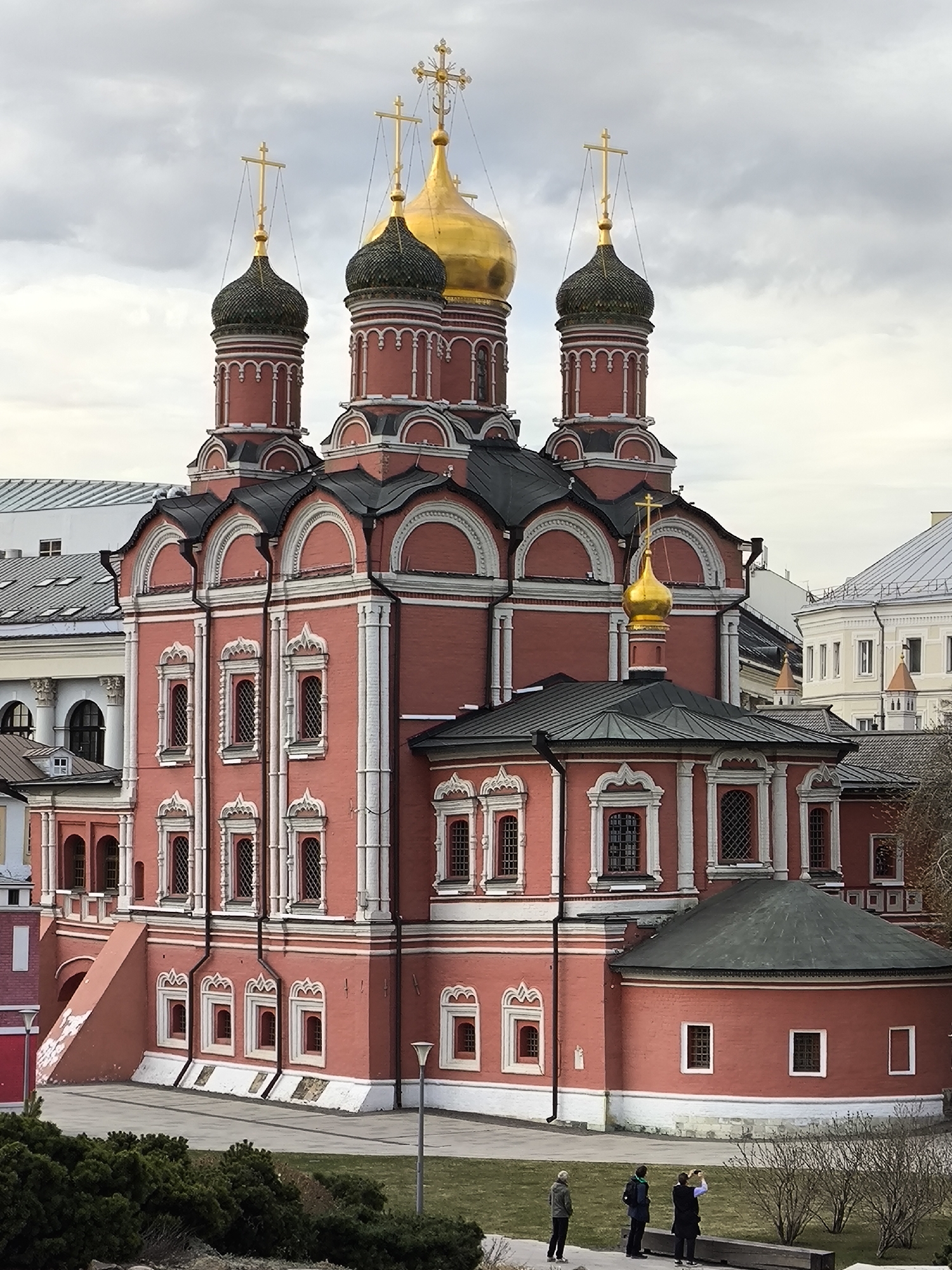 The image presents a view of a Russian Orthodox church in Moscow, Russia. The structure is primarily red and white, featuring multiple levels and traditional onion domes, some of which are gold. Three crosses adorn the top of the building. The building is situated near a patch of greenery. Three people stand on a wooden bench and admire the church. The sky is overcast with grey clouds. The church is surrounded by other buildings.