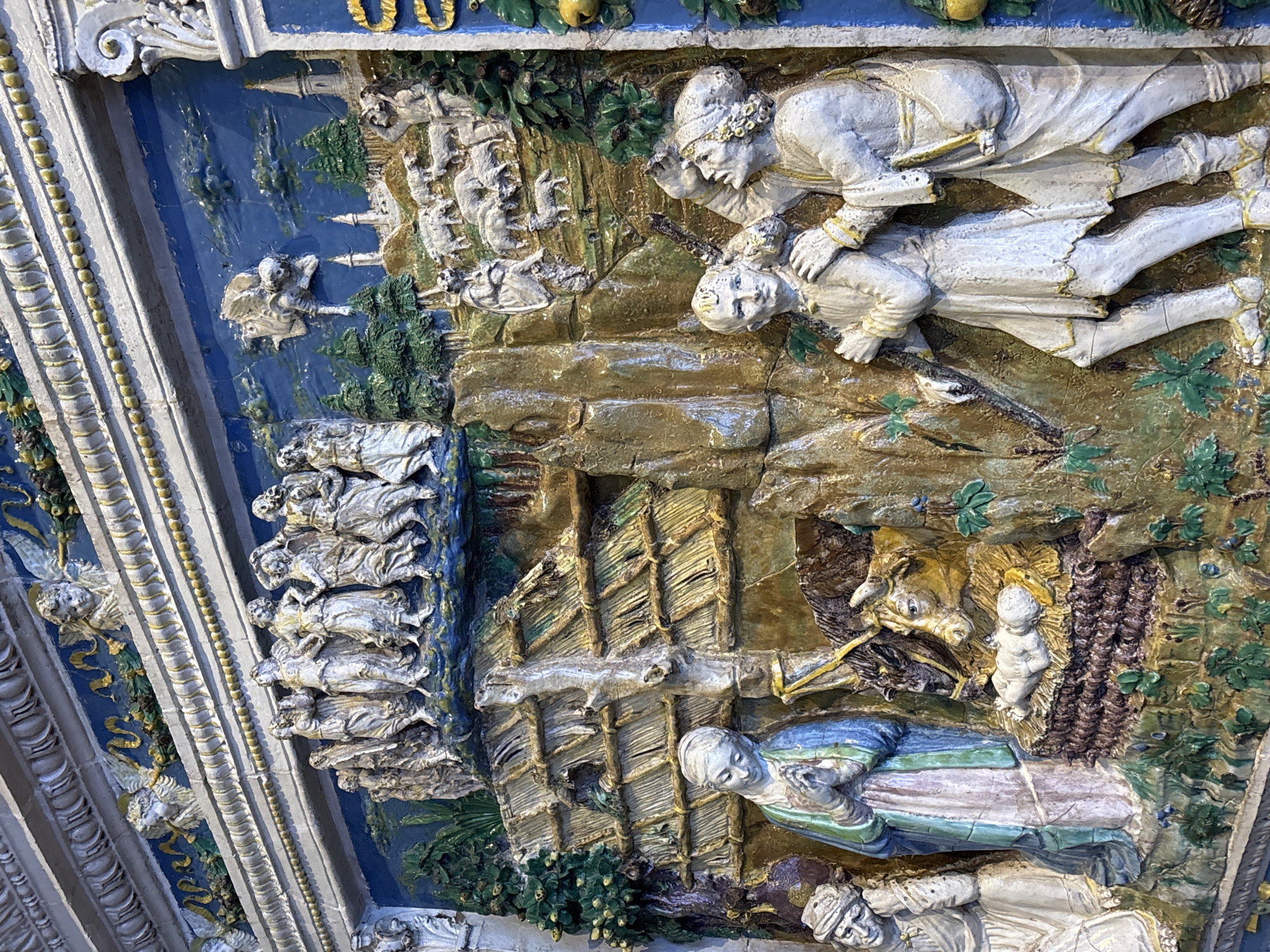 This media file is a close-up, angled shot of a section of a colorful, three-dimensional relief artwork, likely a frieze or decorative panel, mounted on a building or structure. The artwork depicts a narrative scene with multiple figures and elements.
The main subjects are human figures rendered in a stylized, somewhat archaic manner, with pale skin and detailed clothing. In the upper portion, two figures are depicted in motion. One, wearing a helmet adorned with floral motifs, leans forward, seemingly gesturing or interacting with another figure who is lying down. These figures appear to be in a rocky, somewhat barren landscape with green foliage.
Below these figures, the scene shifts to what appears to be a manger or stable setting. A newborn infant, depicted in white, stands in straw. Nearby, a horse or ox, rendered in brown and yellow, is present. A woman, dressed in blue, green, and purple robes, lies down, looking towards the infant. Below her, another figure, possibly a man in a white head covering, is also lying down. The setting includes elements that suggest a rustic structure, possibly made of wood and straw.
The overall color palette is rich, with vibrant blues, greens, yellows, and earthy browns, contrasting with the pale tones of the figures. The relief is highly detailed, with intricate folds in the clothing and textured surfaces for the landscape and structures. The artwork is installed at an angle, giving a sense of depth and perspective. Given the location context of Saint Petersburg, Russia, this artwork may be part of a historic building or an ecclesiastical structure. No visible text is present in the image.