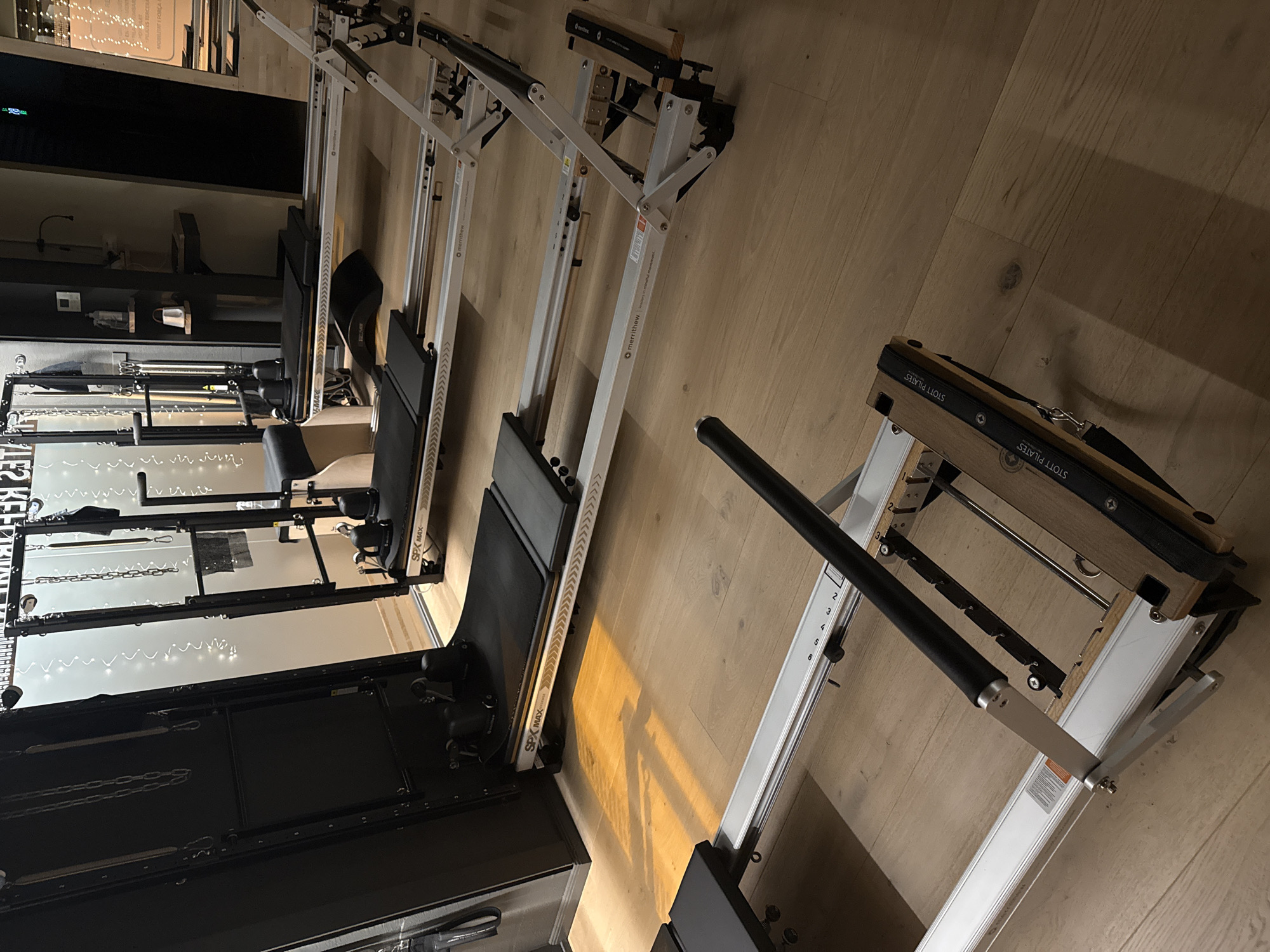 The image shows a line of Pilates machines in what appears to be a studio or home gym setting. The machines are aligned on a wooden floor. Some are black and some are white. The machines are labeled "STOTT PILATES" and "merrithew". A pilates reformer with a footbar is visible. The room contains other fitness-related equipment and shelving. The lighting is warm and ambient. The scene is indoors. There is no visible sign of the city of Barcelona.