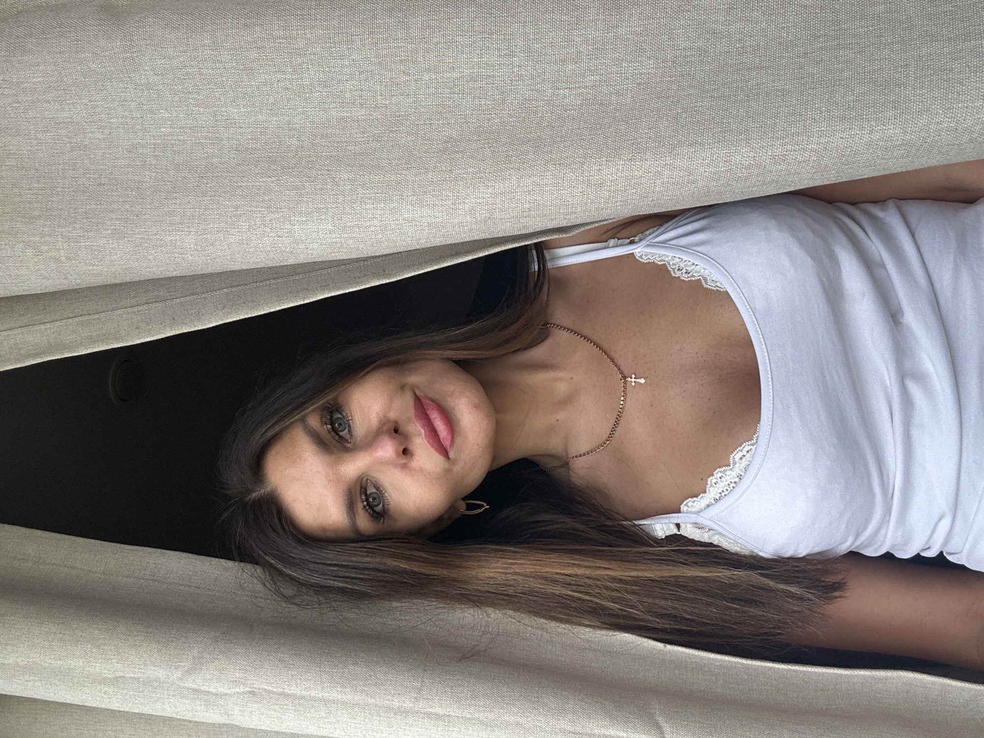 A woman with long brown hair, fair skin, and green eyes peers out from behind a set of light beige curtains. She wears a white tank top with lace trim around the neckline and a delicate gold necklace with a cross pendant. The location is unknown and there is no way to determine the location is in the city of Chaikovsky, Russia from the image.