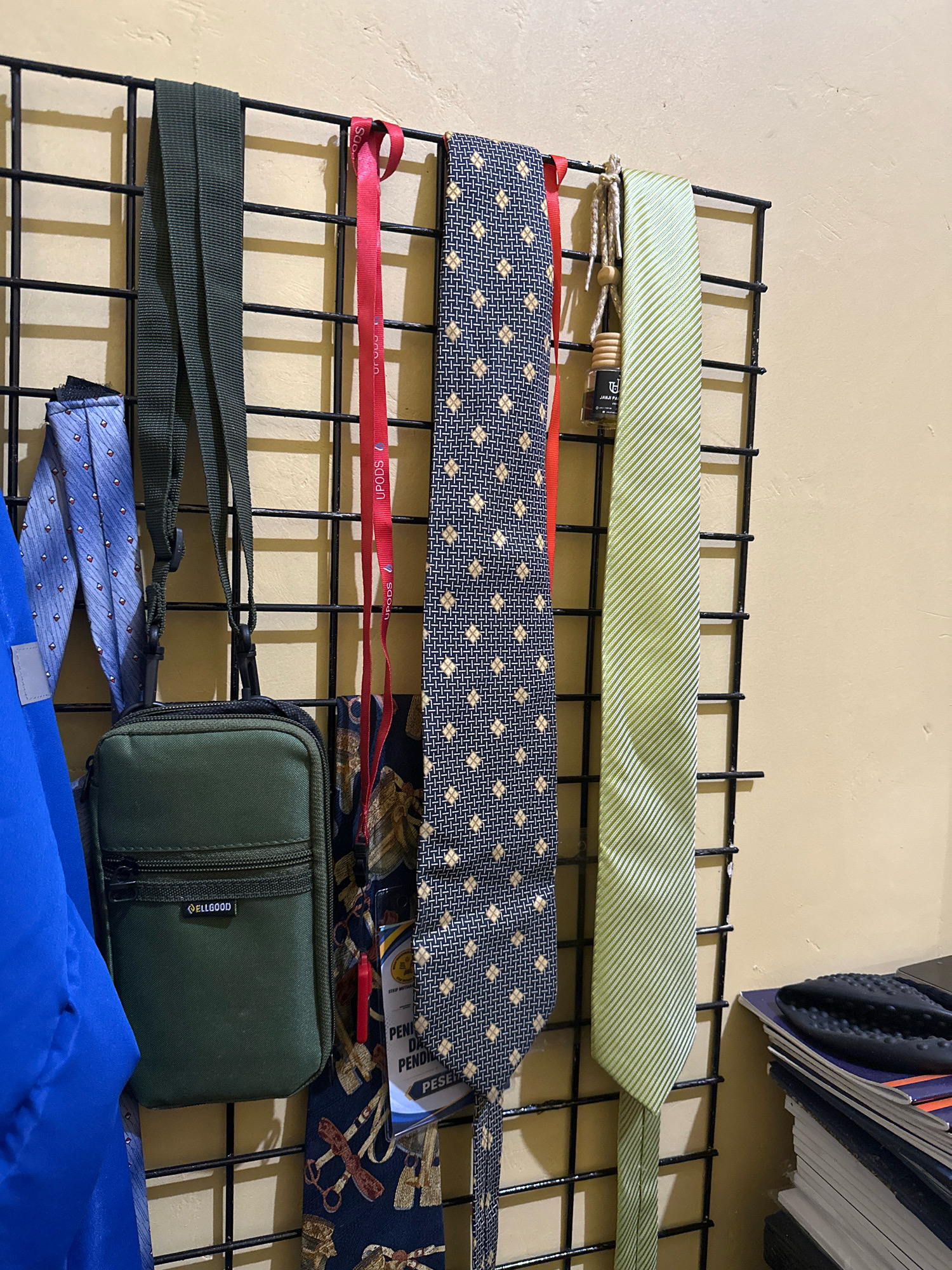 A close-up, eye-level shot captures a variety of ties and a small green pouch hanging on a black metal grid against a beige wall. The grid is positioned in what appears to be a closet or dressing area. The items are arranged from left to right: a blue tie with a diamond pattern, a green pouch with a zipper and a small "ELLGOOD" label, a red ribbon with "UPODS" printed on it, a dark blue tie with a repeating small gold floral pattern, and a light green tie with diagonal stripes. Below these, additional ties with various patterns are visible, along with a small air freshener hanging from twine. To the far right, a stack of books or magazines is partially visible, topped with a black object resembling a shoe insole. The lighting is soft and comes from an unseen source, casting faint shadows on the wall and grid.