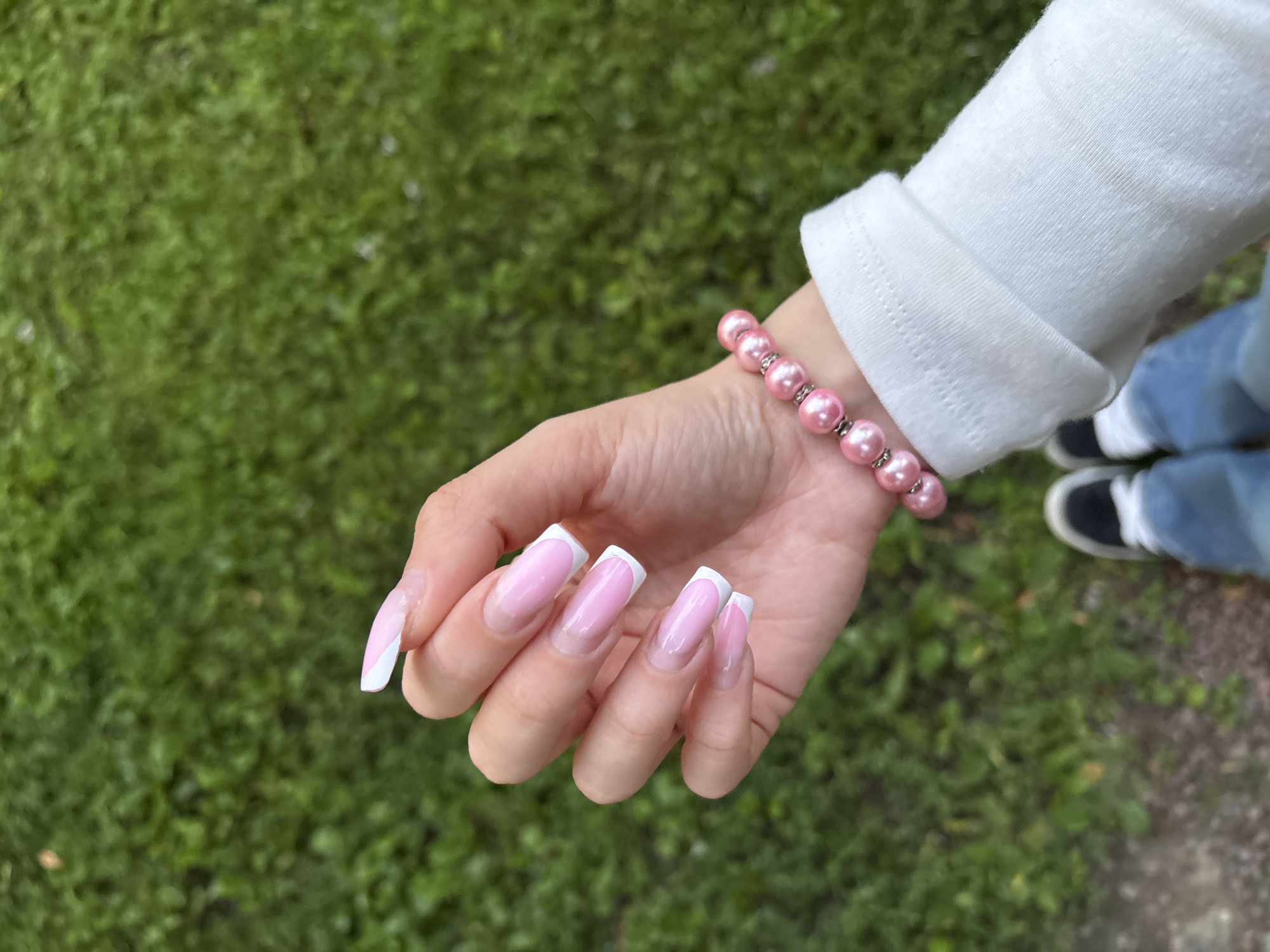 A close-up, overhead shot shows a person's left hand and arm adorned with a pink pearl bracelet and a white long-sleeved shirt. The hand displays a french manicure with pink and white tips. The background is a blurred expanse of green grass, suggesting an outdoor setting. The image does not contain any identifiable features of Pori, Finland.