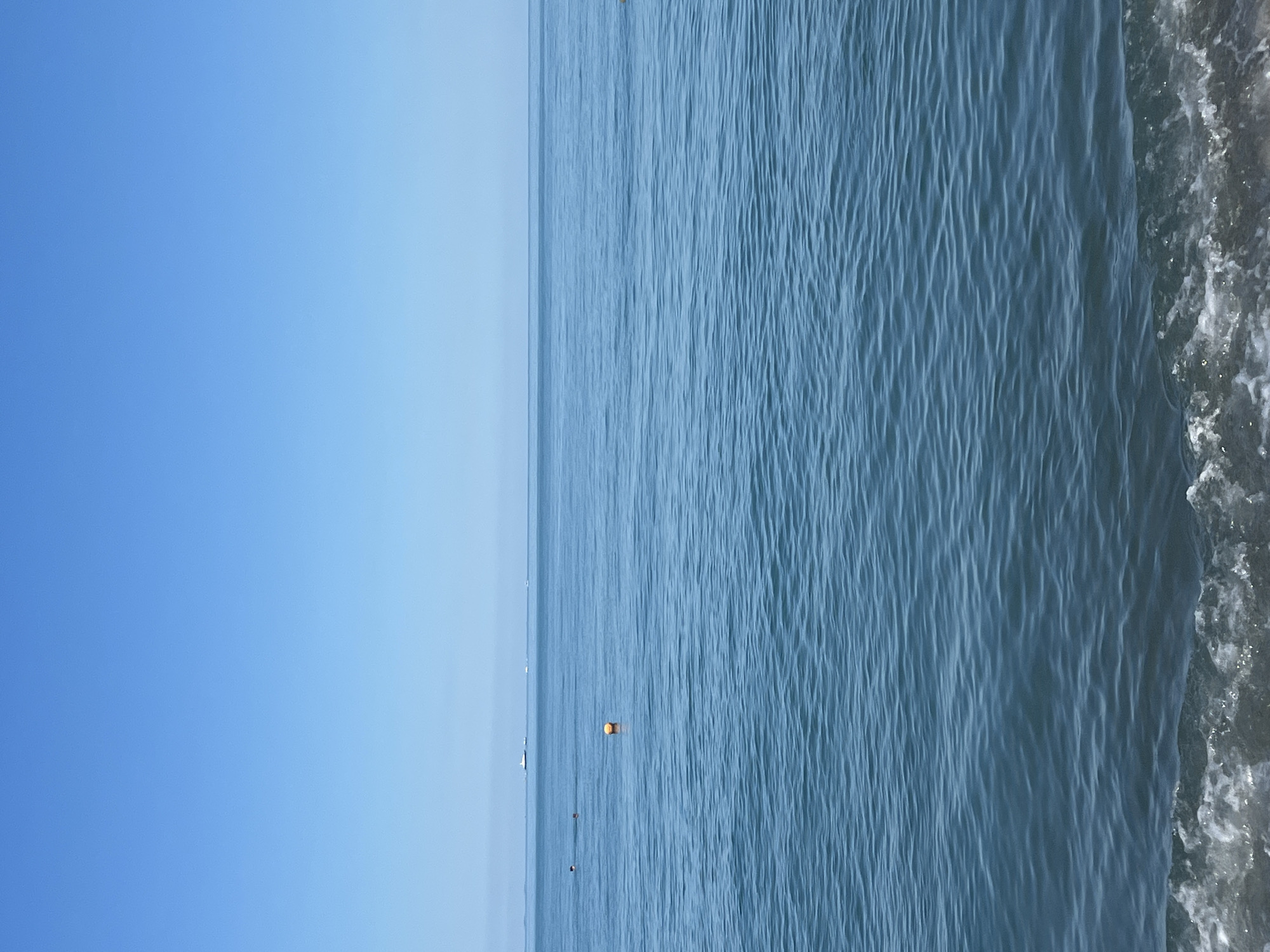 A vertical, eye-level shot captures the tranquil expanse of the sea under a clear, bright blue sky. The horizon is a sharp line separating the water from the sky, with a faint, distant outline of a large white boat and smaller vessels on the left side. In the middle distance, a singular orange buoy bobs gently on the surface of the water. To the left of the buoy, two figures are visible in the water, appearing to swim or tread water. The sea itself is a deep, serene blue, rippled by gentle waves that are more pronounced in the foreground, where they break into white foam against a dark, indistinct shore. The scene suggests a peaceful day at the beach, likely in the city of Sochi, Russia, known for its Black Sea coastline.