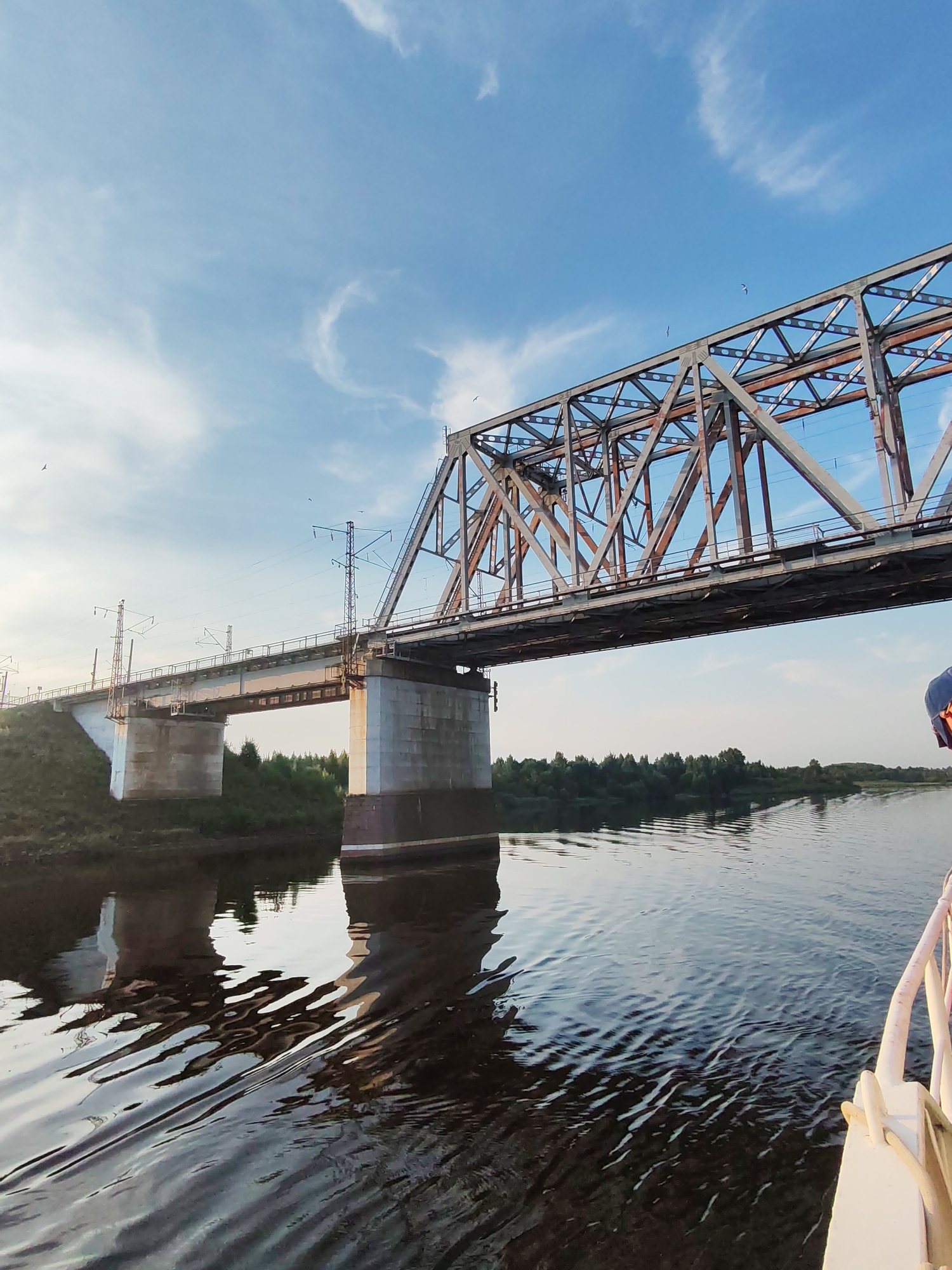 The image shows a large, metal railway bridge spanning a wide river under a bright blue sky with scattered white clouds. The bridge has a distinctive lattice structure, and electric power lines for trains are visible overhead. The river is dark and reflective, with ripples on the surface. Green trees line the riverbank on the far side, and a grassy slope is visible on the near bank. Part of a white railing, possibly from a boat, is visible in the lower right corner of the frame, suggesting the photo was taken from the water. The location is the city of Tarkanovo, Russia.