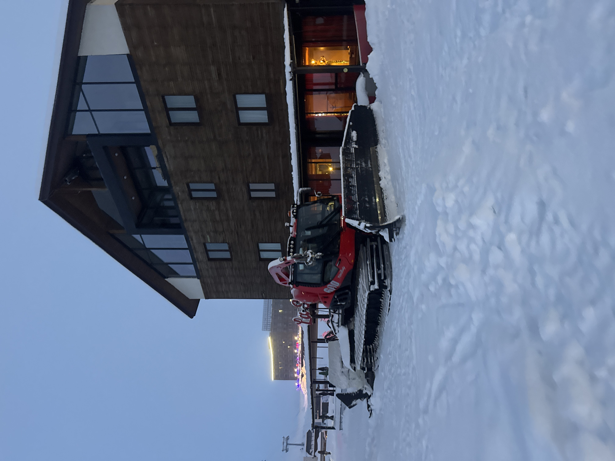 The image depicts a snow-covered scene in front of a modern, multi-story building with a gable roof. The building has dark wood siding and large windows. A red snow groomer is parked in the foreground, partially obscured by snow. In the background, a distant building is visible with festive lighting. The sky is a pale blue, suggesting it is early morning or dusk. The overall environment appears to be a ski resort or mountain village during winter. The location is Unknown, Unknown.