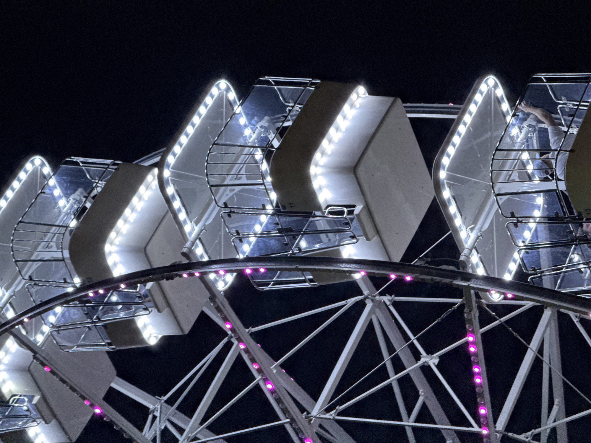 The image is a close-up shot of a Ferris wheel at night in Budva, Montenegro. The Ferris wheel is illuminated with white and pink lights. Several gondolas are visible, each with glass panels and metal railings. In one of the gondolas, a person wearing a light blue cap and white shirt is visible, holding a blue object and looking upwards. The background is a dark, night sky. The overall impression is that of a festive amusement park scene.