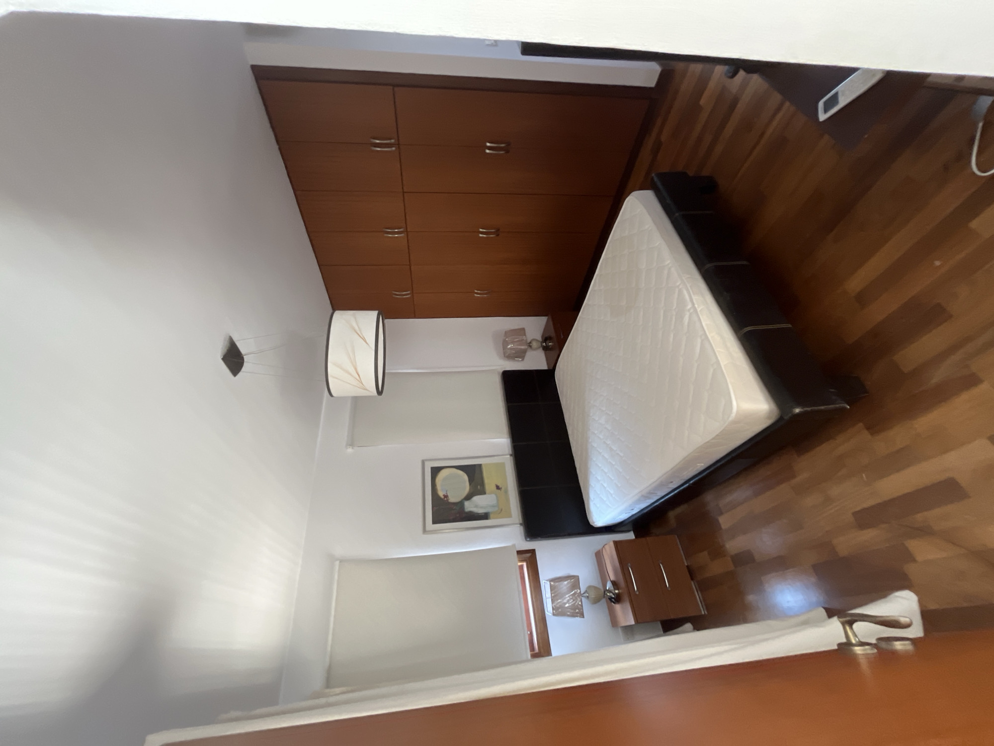 The image shows a bedroom in Aphrodite Hills, Cyprus. The room has a queen-sized bed with a black headboard and a white mattress. On the left side of the bed, there is a bedside table with a lamp. In the background, there is a large wooden wardrobe with multiple doors. A painting hangs on the wall above the bedside table. The floor is made of polished wood. The room appears to be well-lit, with natural light coming from a window covered by a white blind.